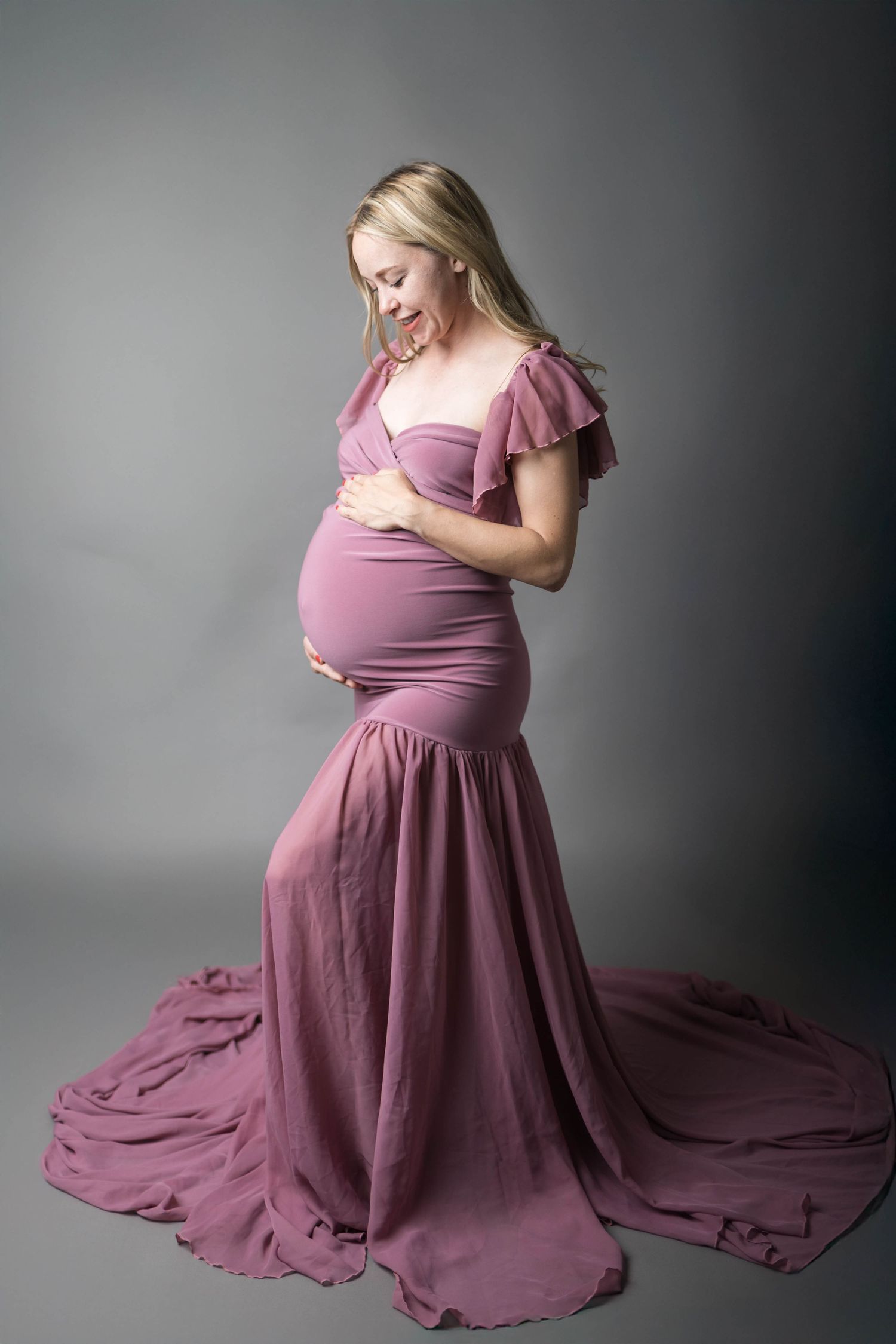 Pregnant woman in pink mauve maternity gown cradling baby bump, smiling down, studio portrait on gray background.