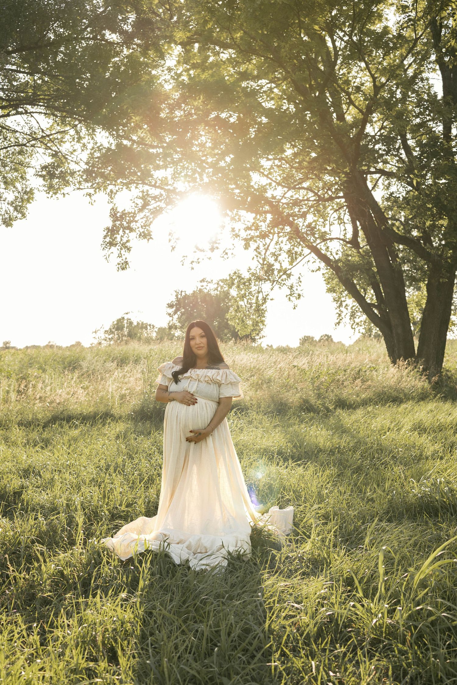 Pregnant woman in white off-shoulder gown standing in sunlit meadow with trees.
