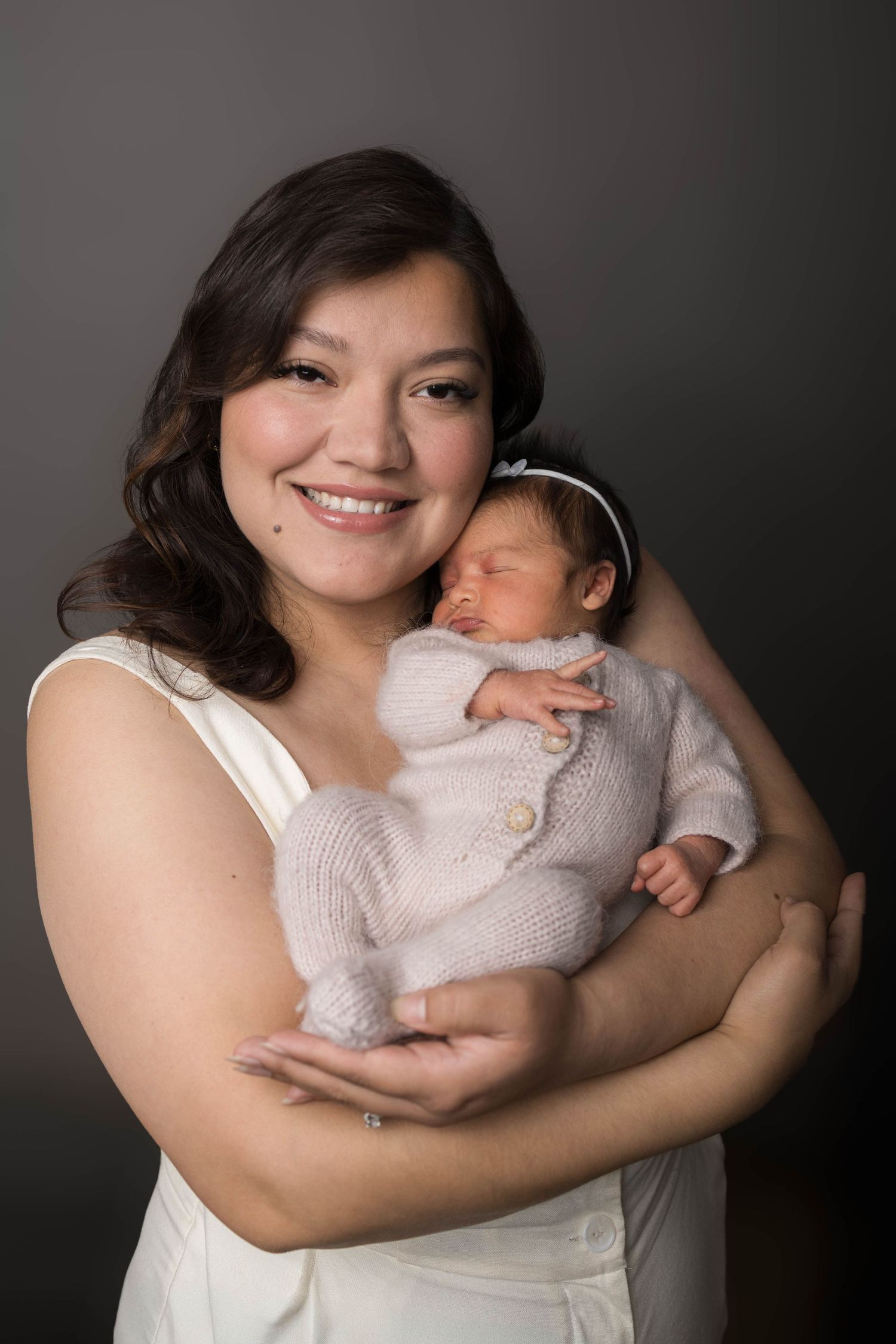 Smiling mother in white dress holds sleeping newborn baby dressed in pink knit outfit with headband.