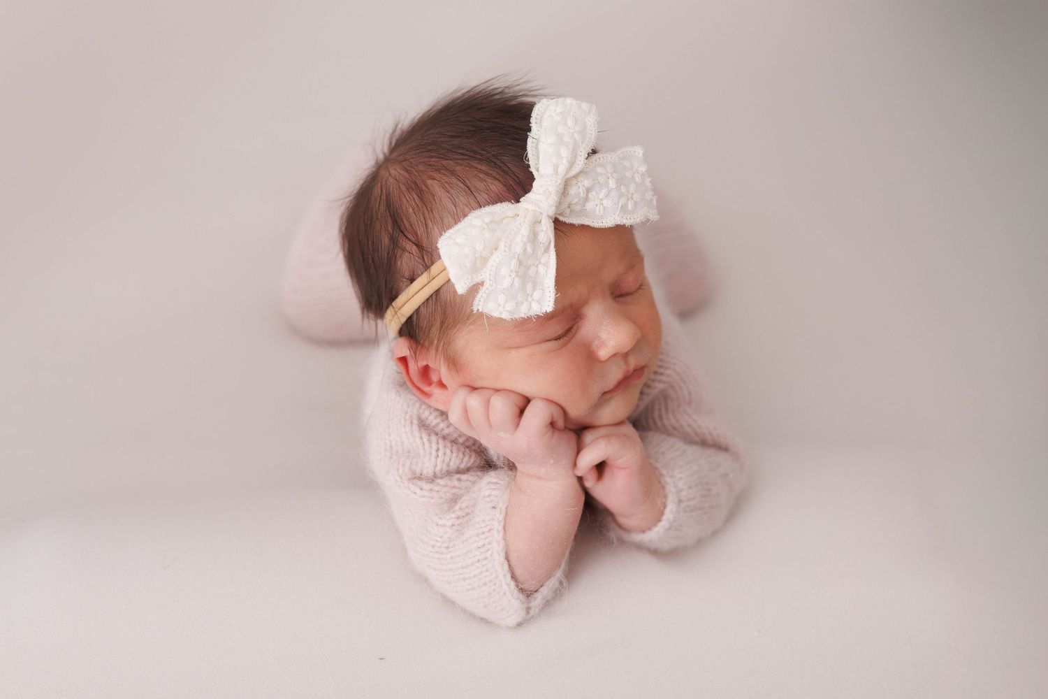 Newborn baby in pink outfit with white bow headband sleeping peacefully on soft backdrop.