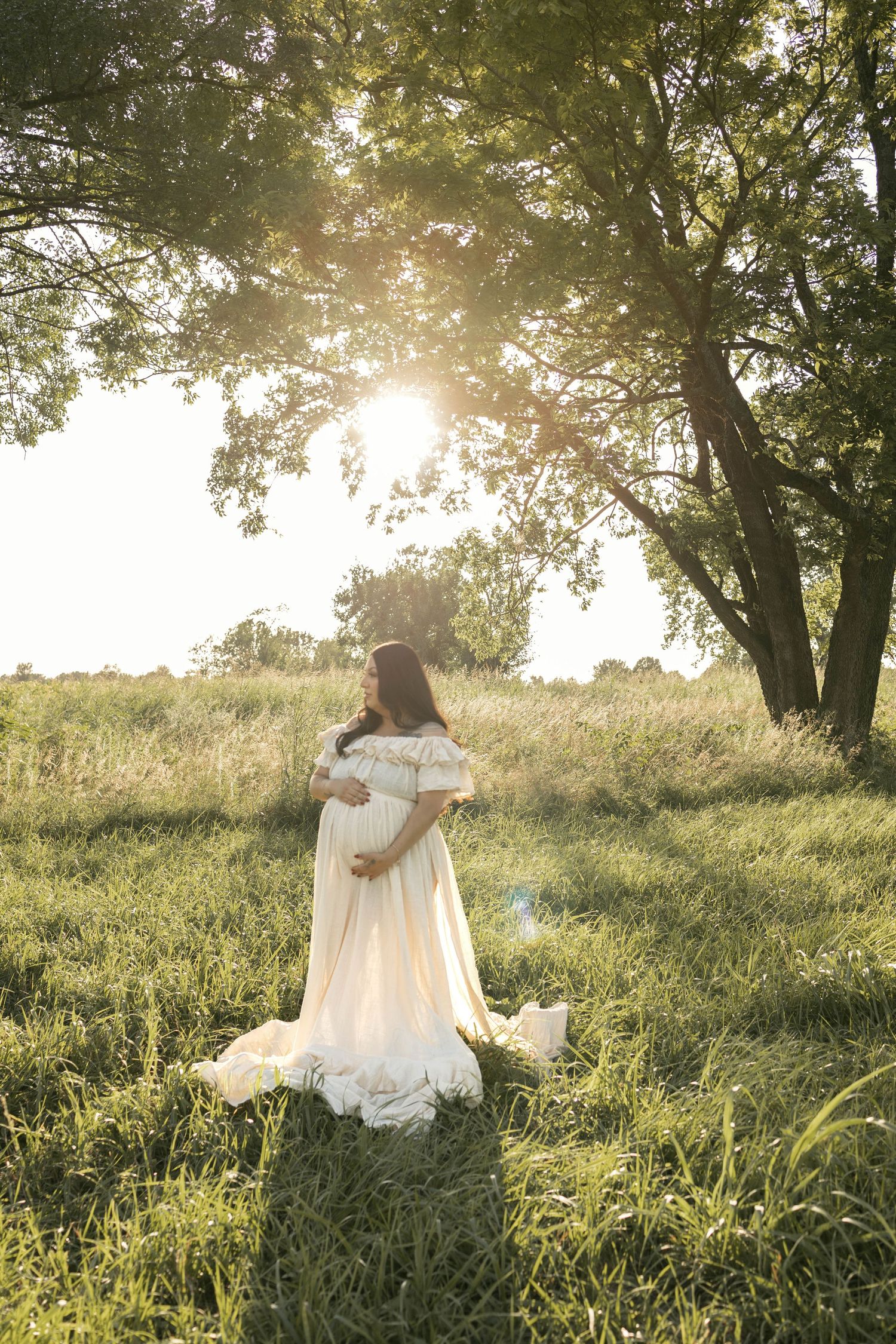 Pregnant woman in flowing white gown viewed from behind in sunlit green field.