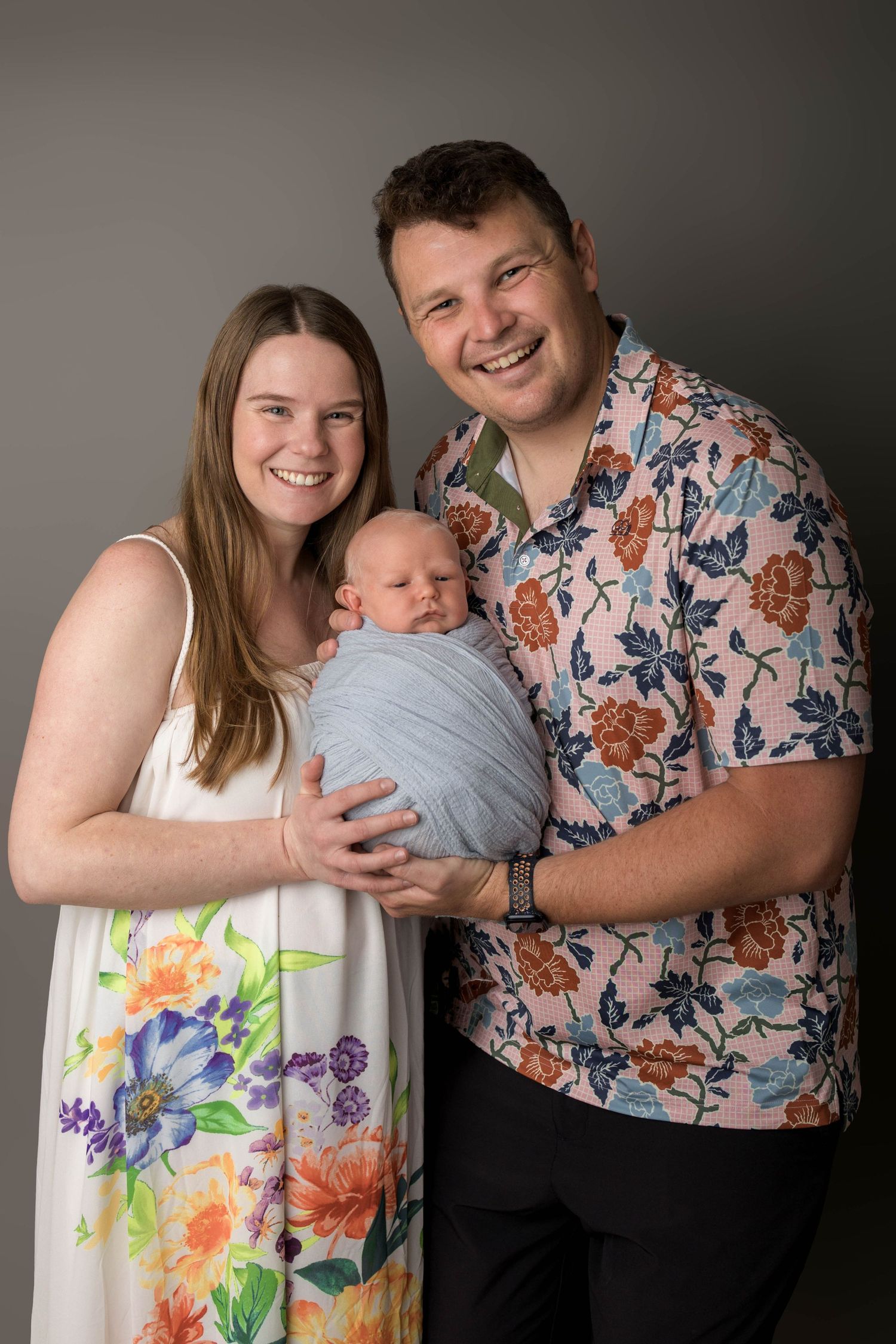 Happy couple smiling and holding their newborn baby wrapped in a grey swaddle, wearing floral outfits.