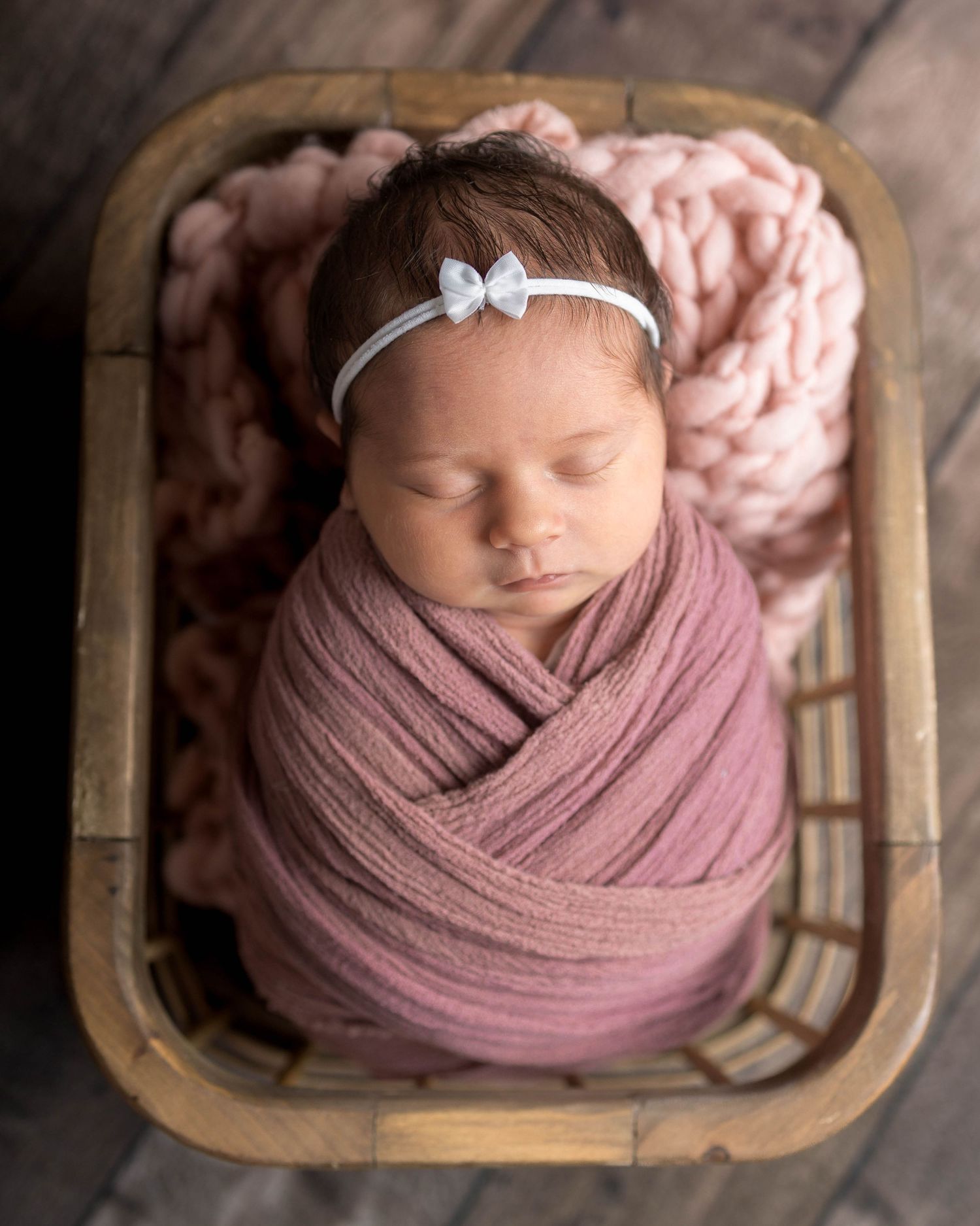 Newborn baby girl swaddled in pink wrap with white bow headband, sleeping peacefully in a rustic wooden basket.