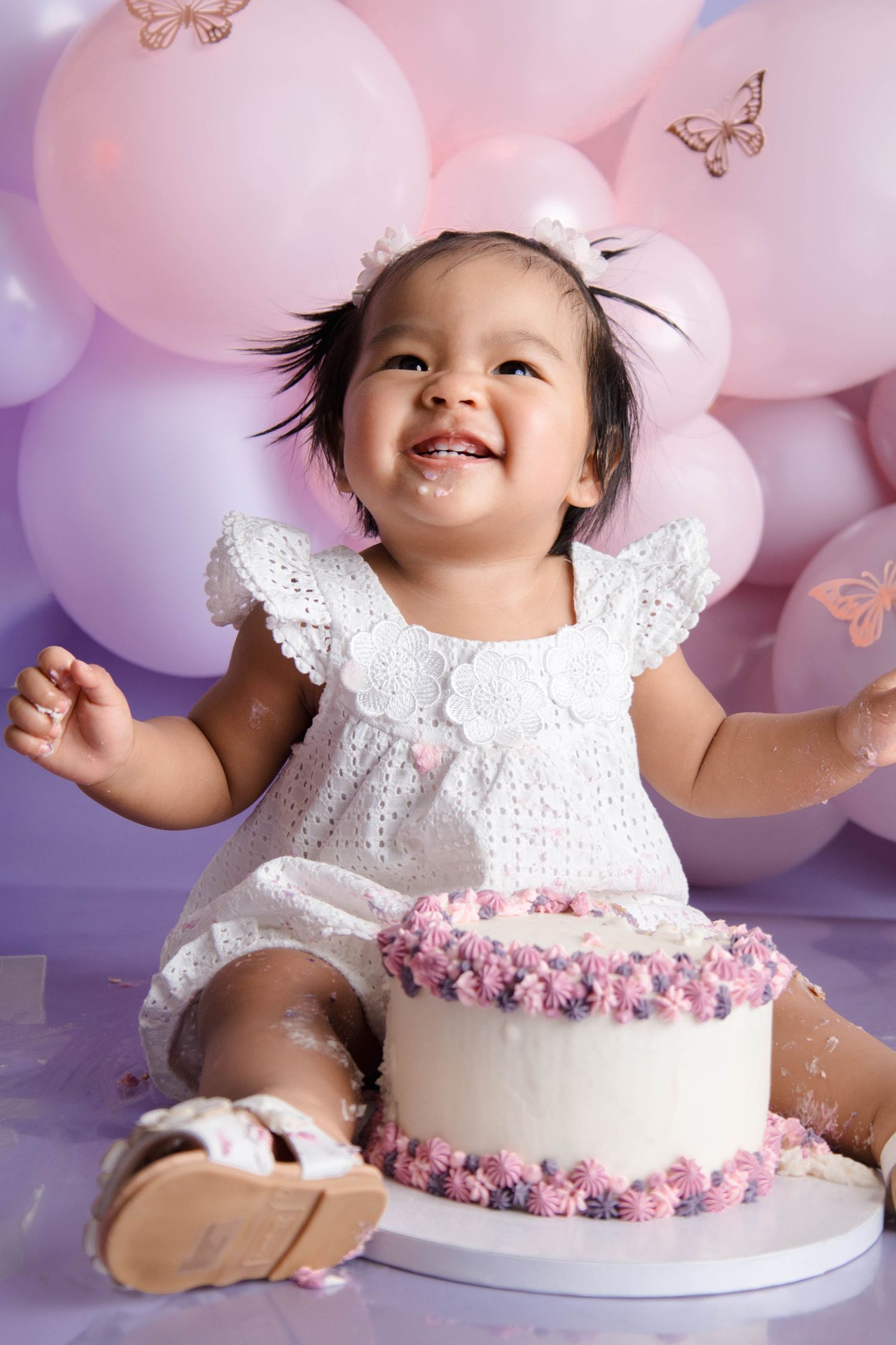 A baby in a white lace dress smiles and plays with pink and purple balloons during a cake smash photo session.