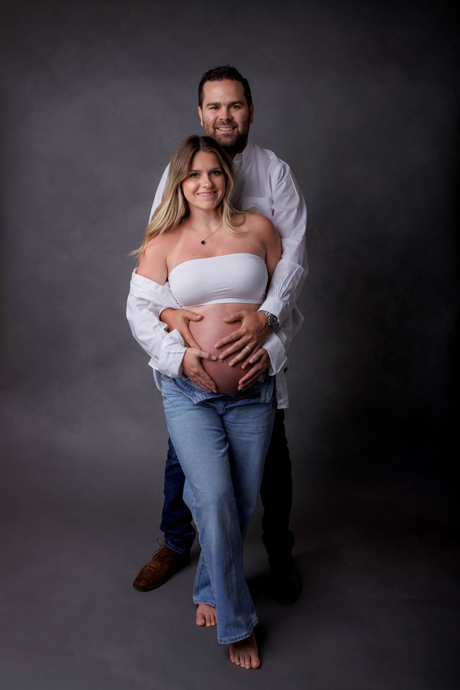 Couple posing for maternity photo, man embracing pregnant woman from behind, both smiling, studio grey background.