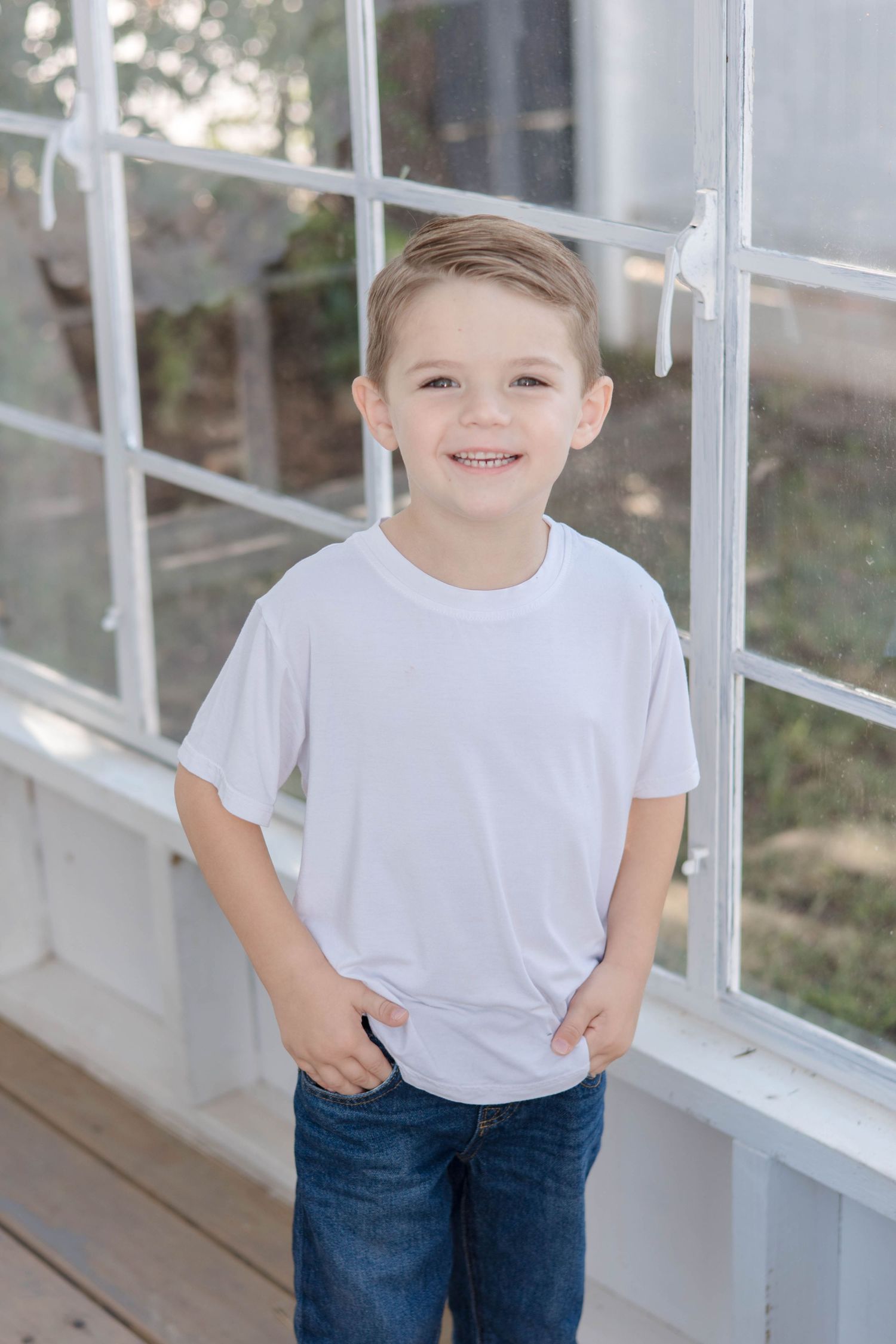 A child in a white t-shirt and jeans stands by a window pane in bright natural lighting.