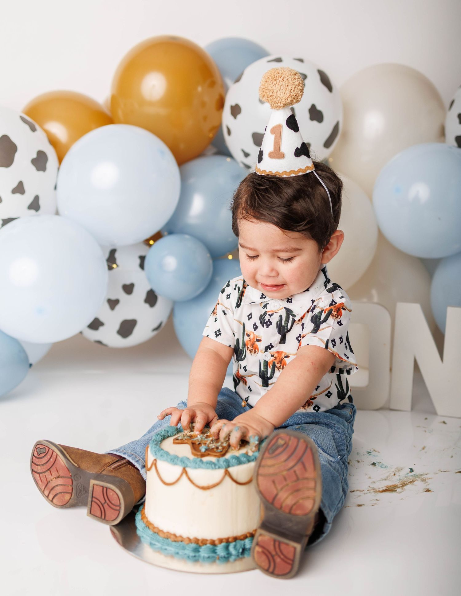 Baby boy in birthday hat smashes first cake surrounded by cow-print and blue balloons at western-themed party.