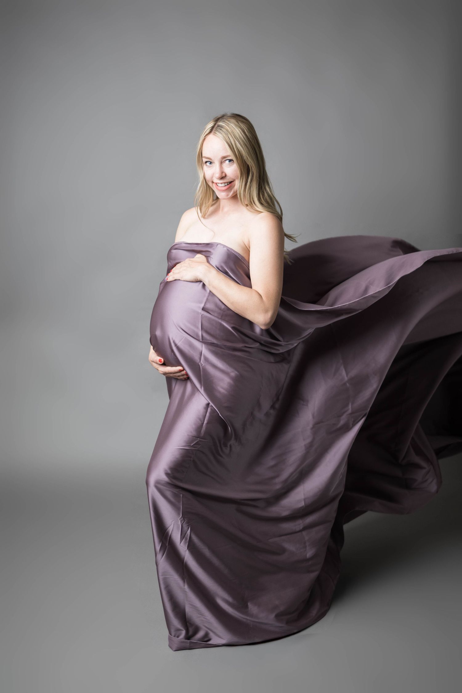 Pregnant woman draped in flowing purple satin fabric, smiling gracefully against a gray studio background.