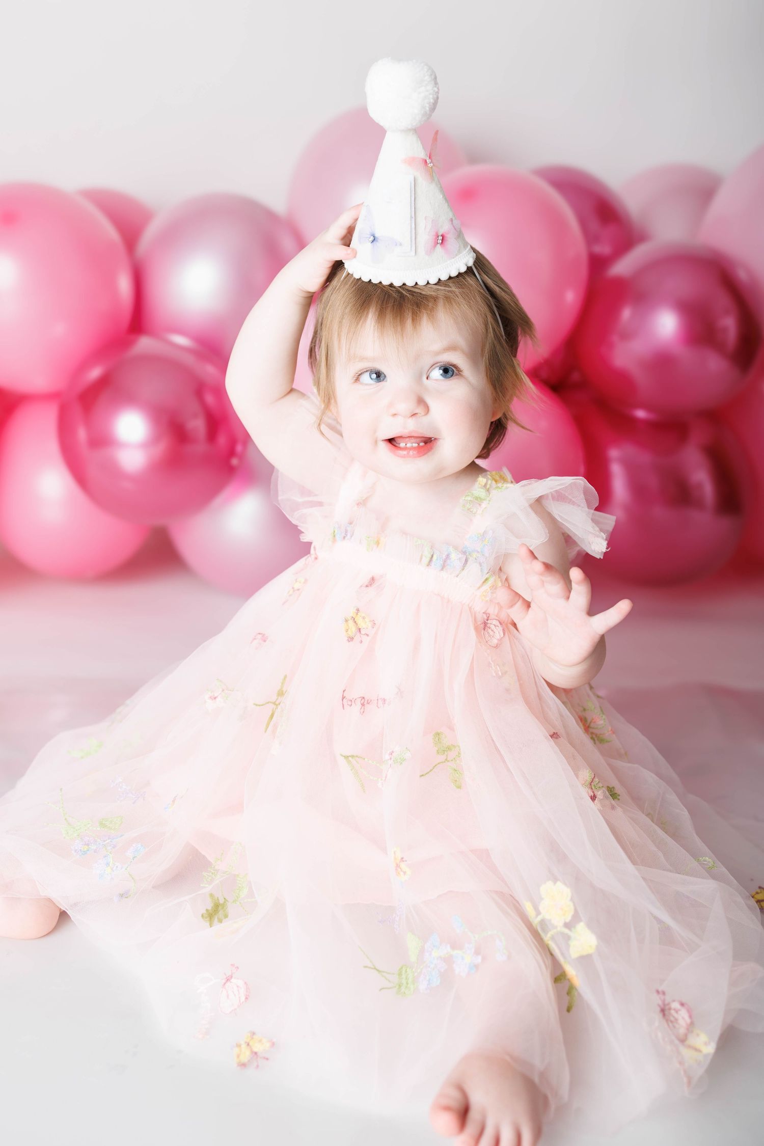 Smiling baby girl in pink tulle dress and party hat sitting in front of pink balloons at her first birthday celebration.