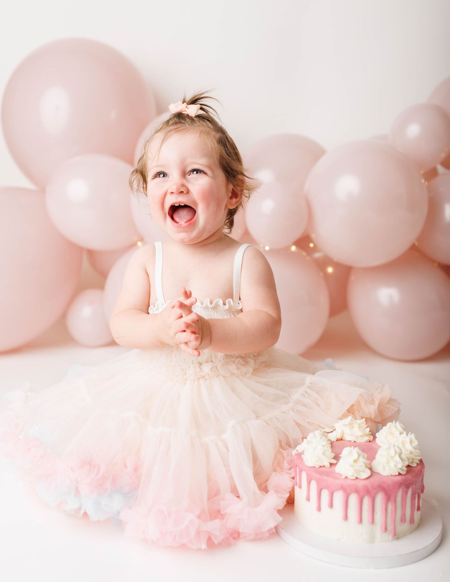 Toddler girl in pink tutu dress laughing at first birthday cake smash session with pink balloon backdrop.