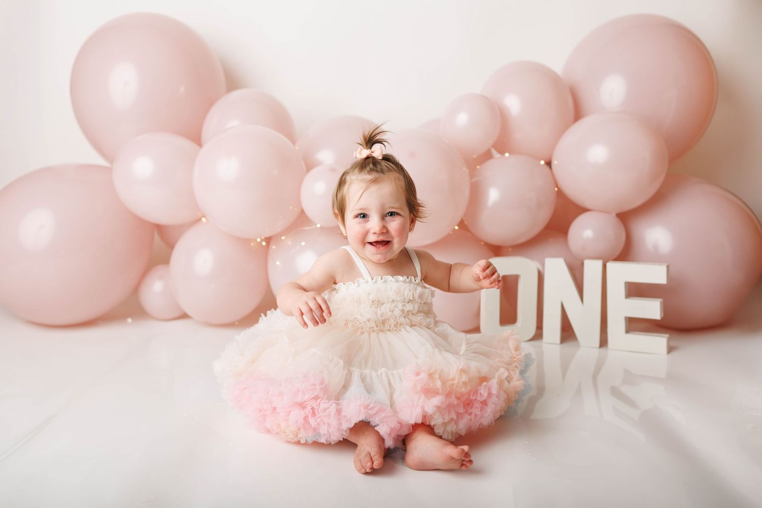Baby girl in pink tutu dress sits smiling before pink balloon arch with ONE sign at her first birthday cake smash.