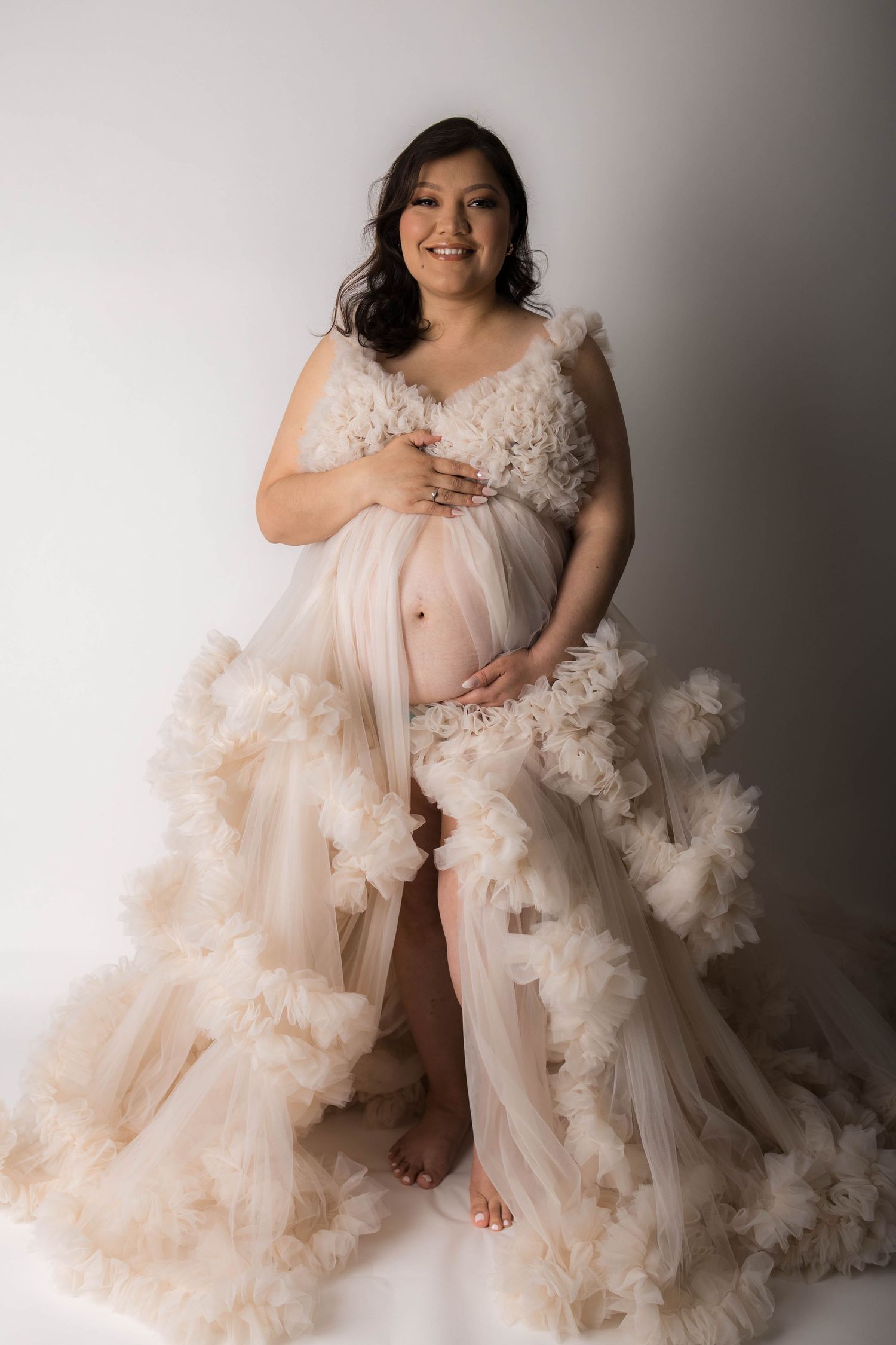 Smiling pregnant woman poses in a flowing cream ruffle tulle maternity gown against a white studio background.