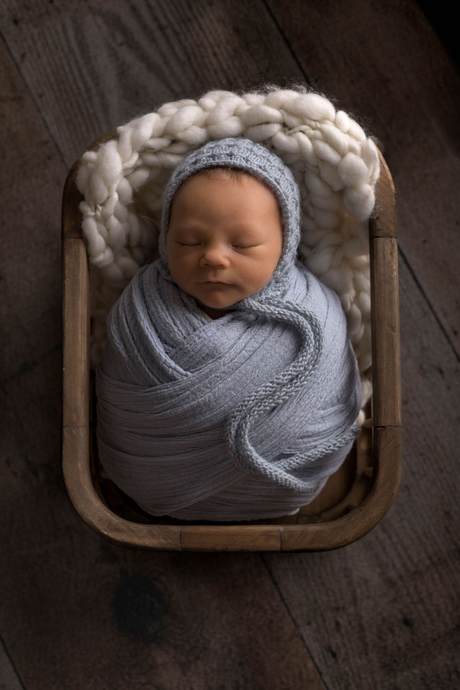 Newborn baby swaddled in soft gray knit blanket sleeping peacefully in wooden prop bed.
