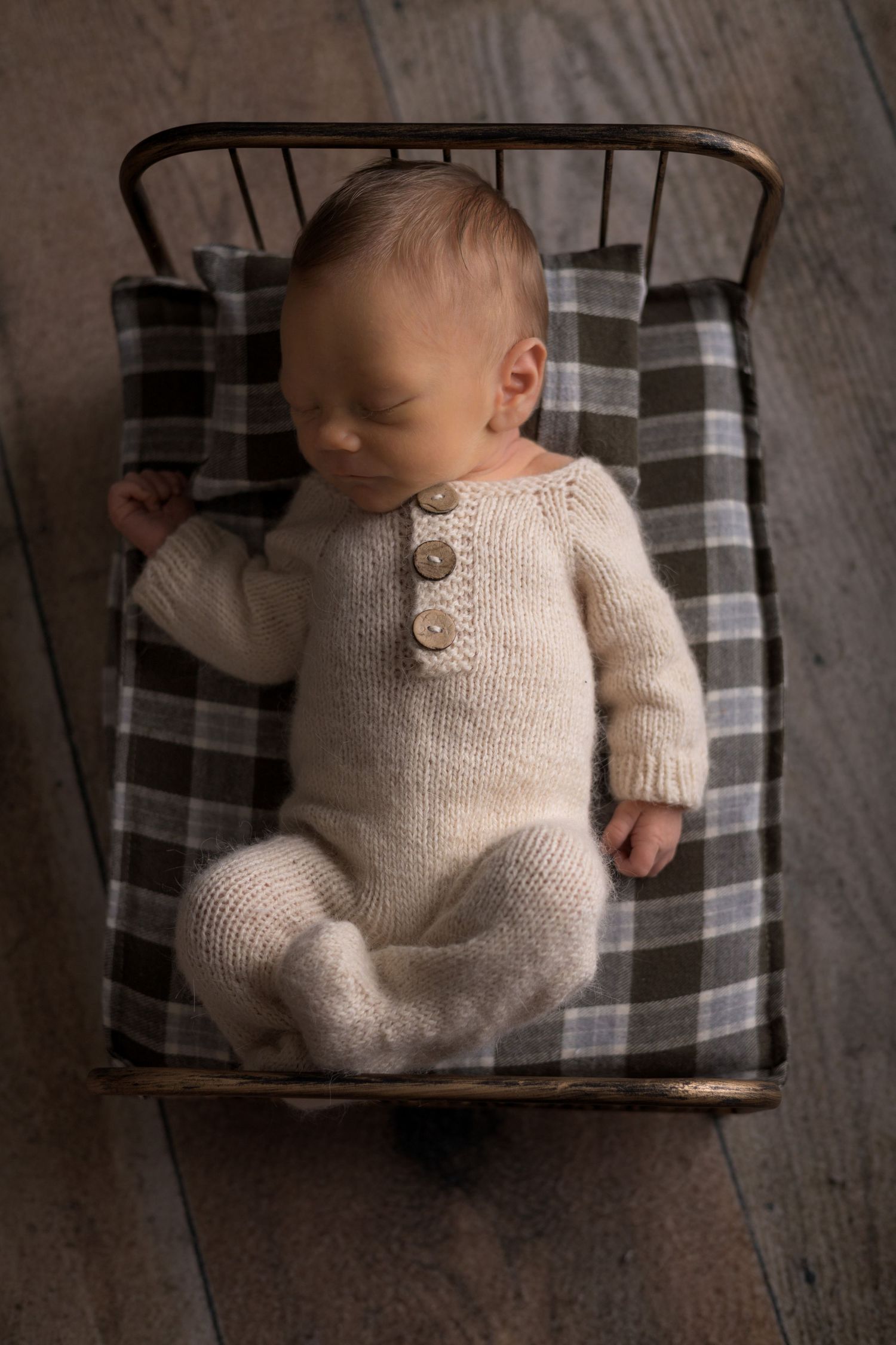 Newborn baby wearing a cream knit sweater lies peacefully on a plaid blanket in a rustic wooden setting.