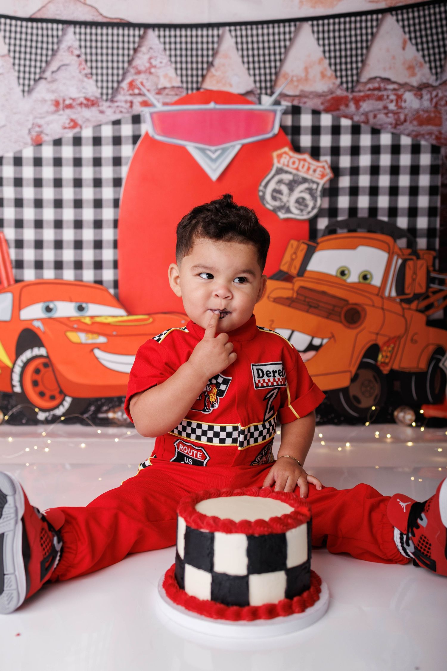 Toddler in red Cars outfit celebrates first birthday with checkered cake in front of Lightning McQueen backdrop.