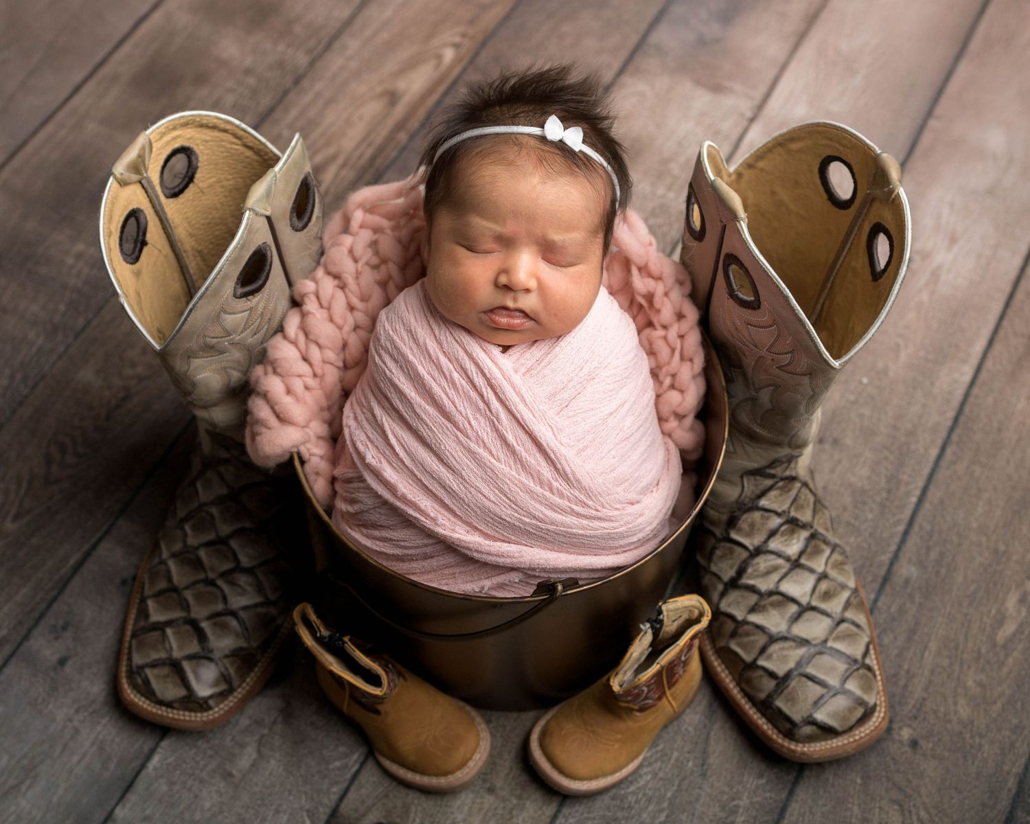 Newborn baby wrapped in pink swaddle sleeping in a bucket surrounded by cowboy boots on a wooden floor.