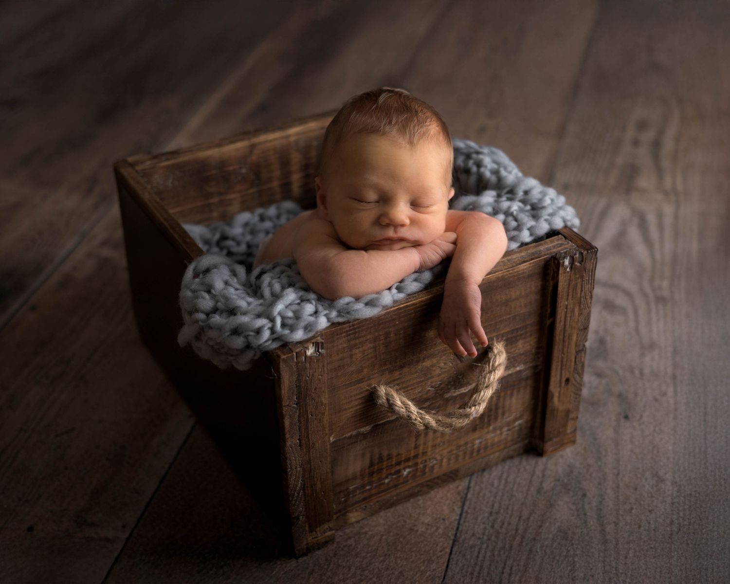 Sleeping newborn rests peacefully in rustic wooden box lined with soft gray blanket on hardwood floor.