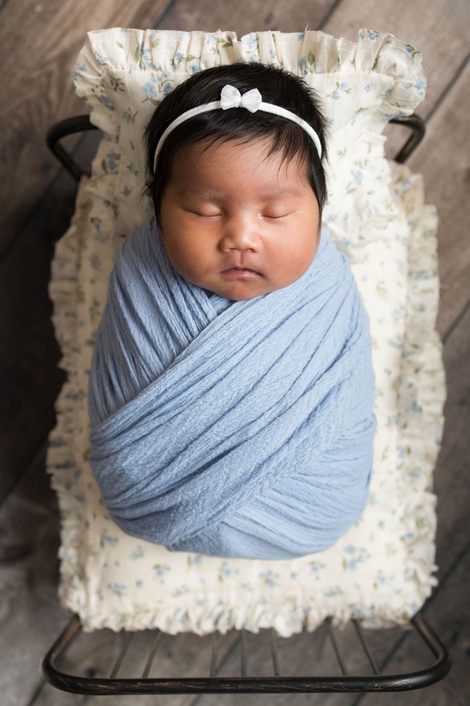 Newborn baby girl swaddled in blue wrap with white bow headband sleeping peacefully in a small vintage metal crib.