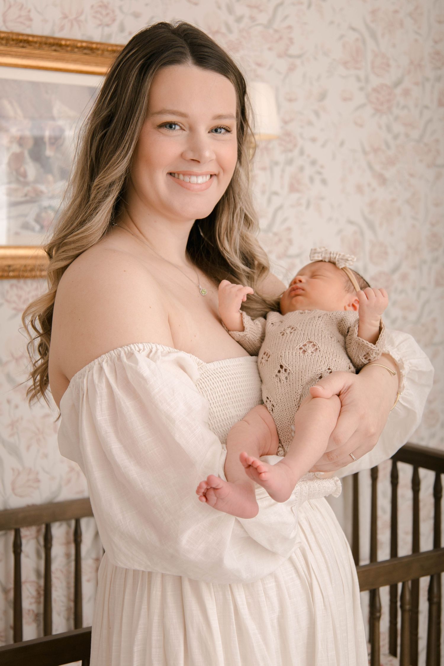 A woman in a white off-shoulder dress holds a sleeping newborn baby against floral wallpaper background.