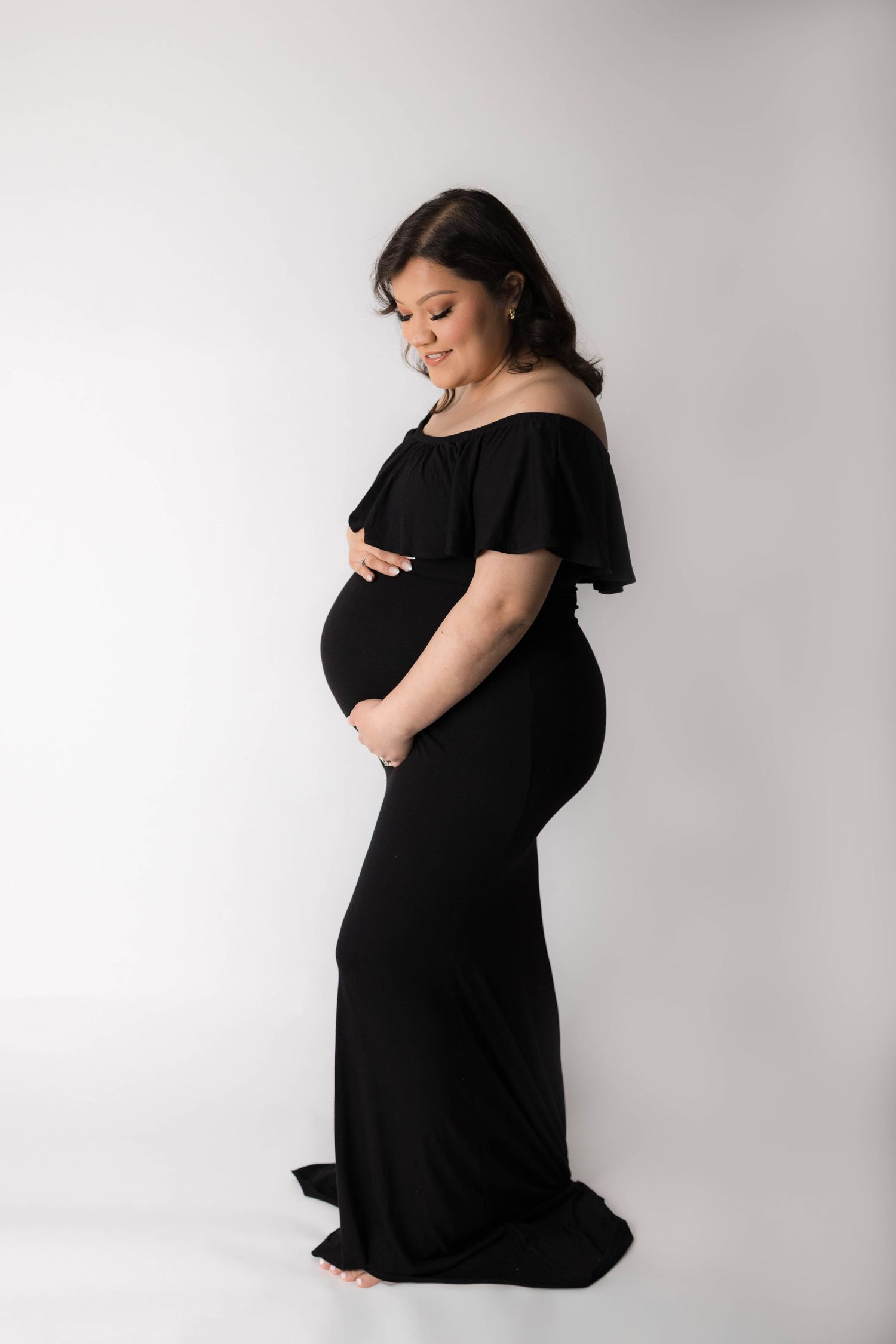 Pregnant woman in elegant black off-shoulder gown cradling baby bump, smiling softly in professional maternity photo.