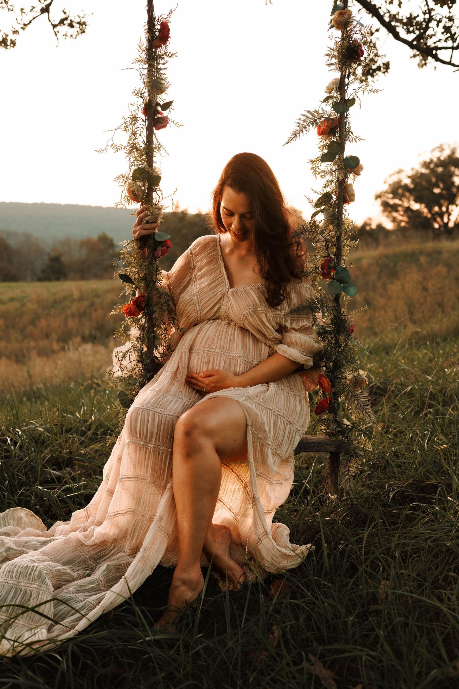 Ethereal maternity photos in field at sunset with floral swing and flowing white dress.
