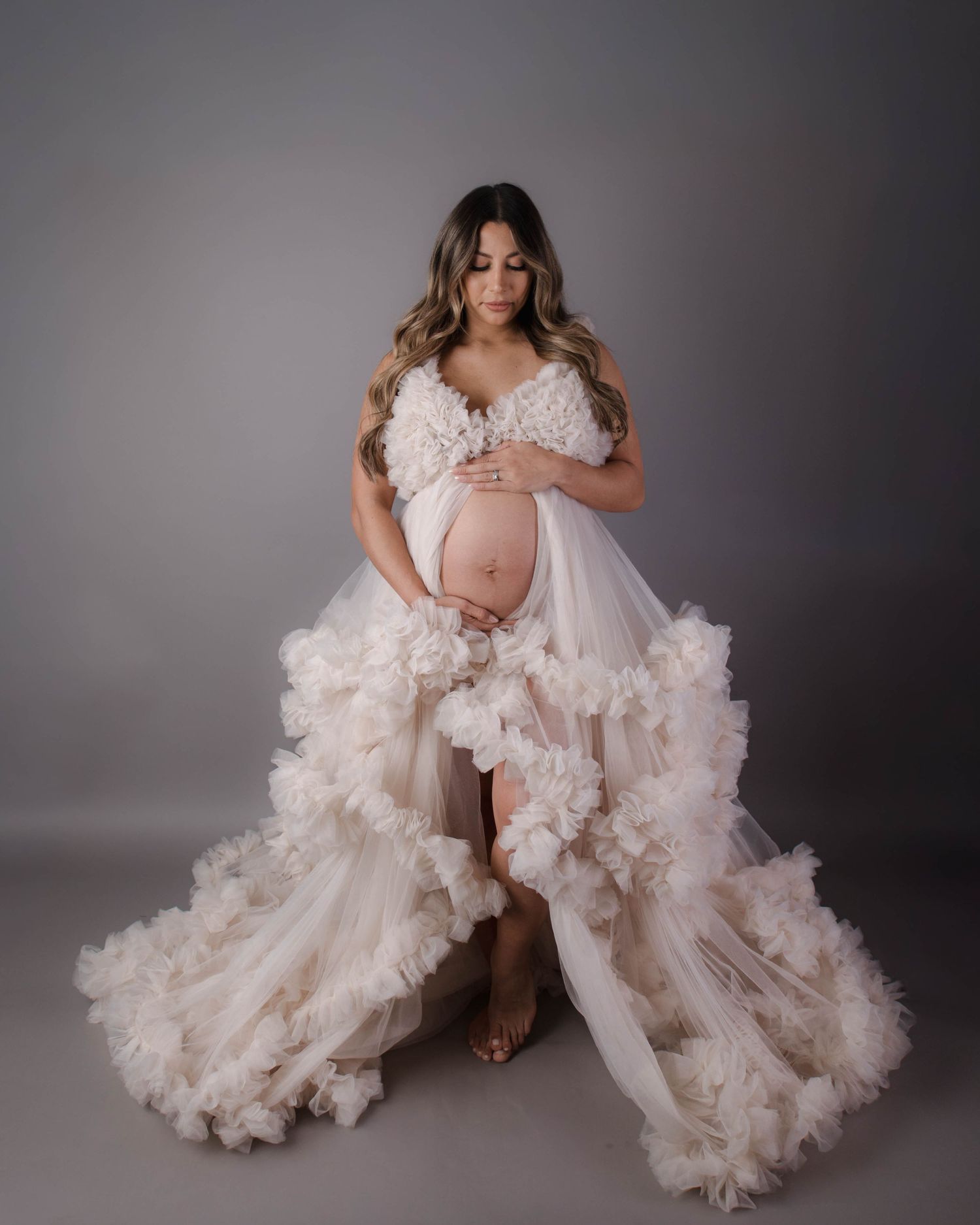 Pregnant woman in a flowing ruffle tulle maternity gown cradling her baby bump during a studio photoshoot.