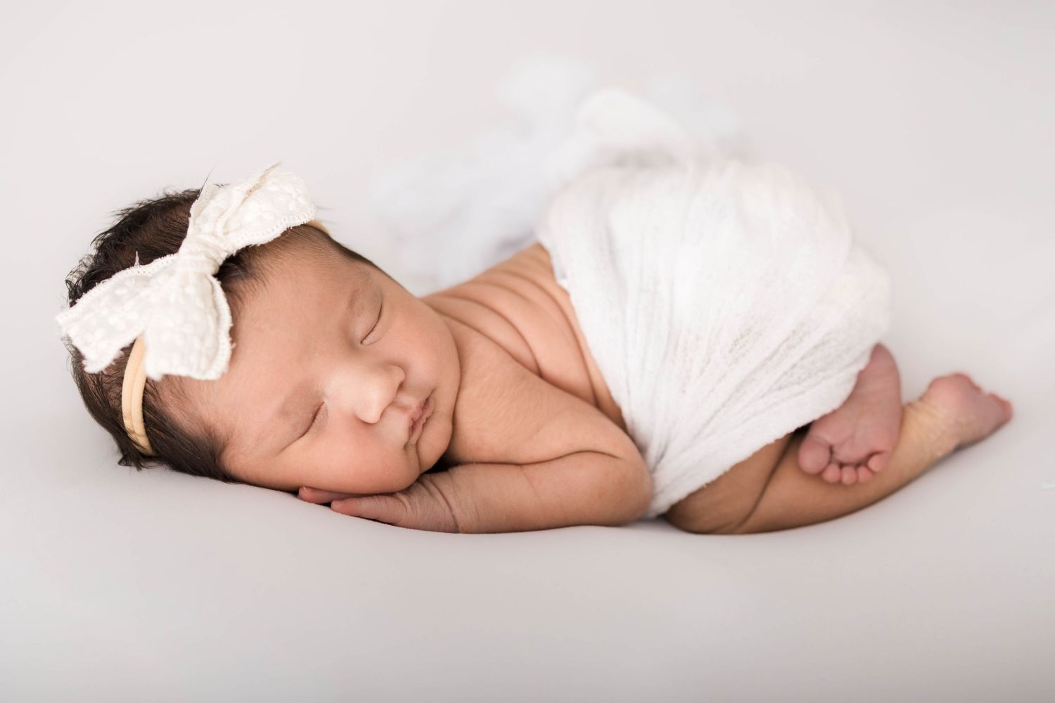 Newborn baby girl sleeping peacefully wrapped in white fabric with a lace bow headband on a white background.