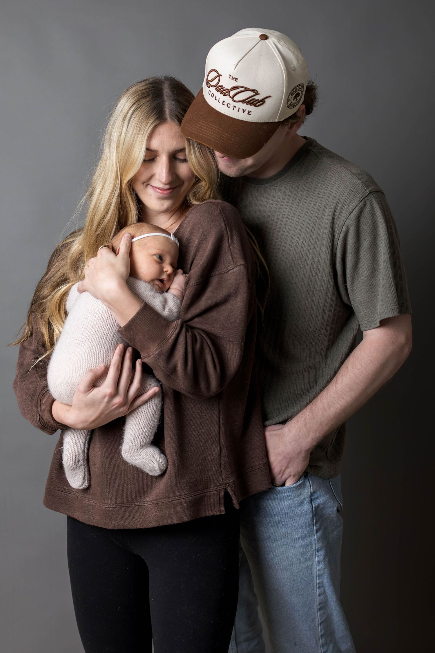 A young couple in casual attire tenderly holds their sleeping newborn baby during a studio portrait session.