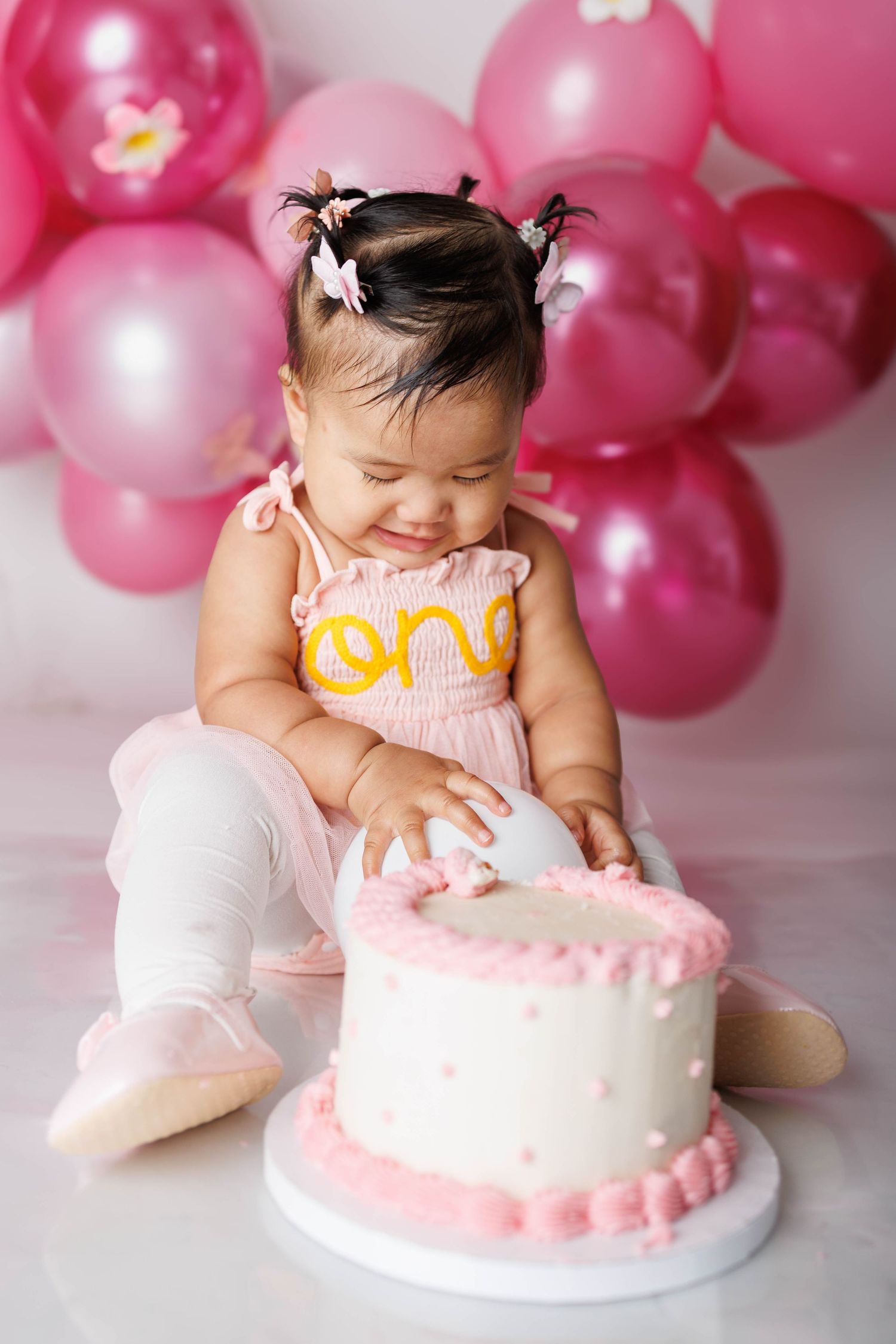 Baby girl in pink ONE dress smashing her first birthday cake surrounded by pink balloons.