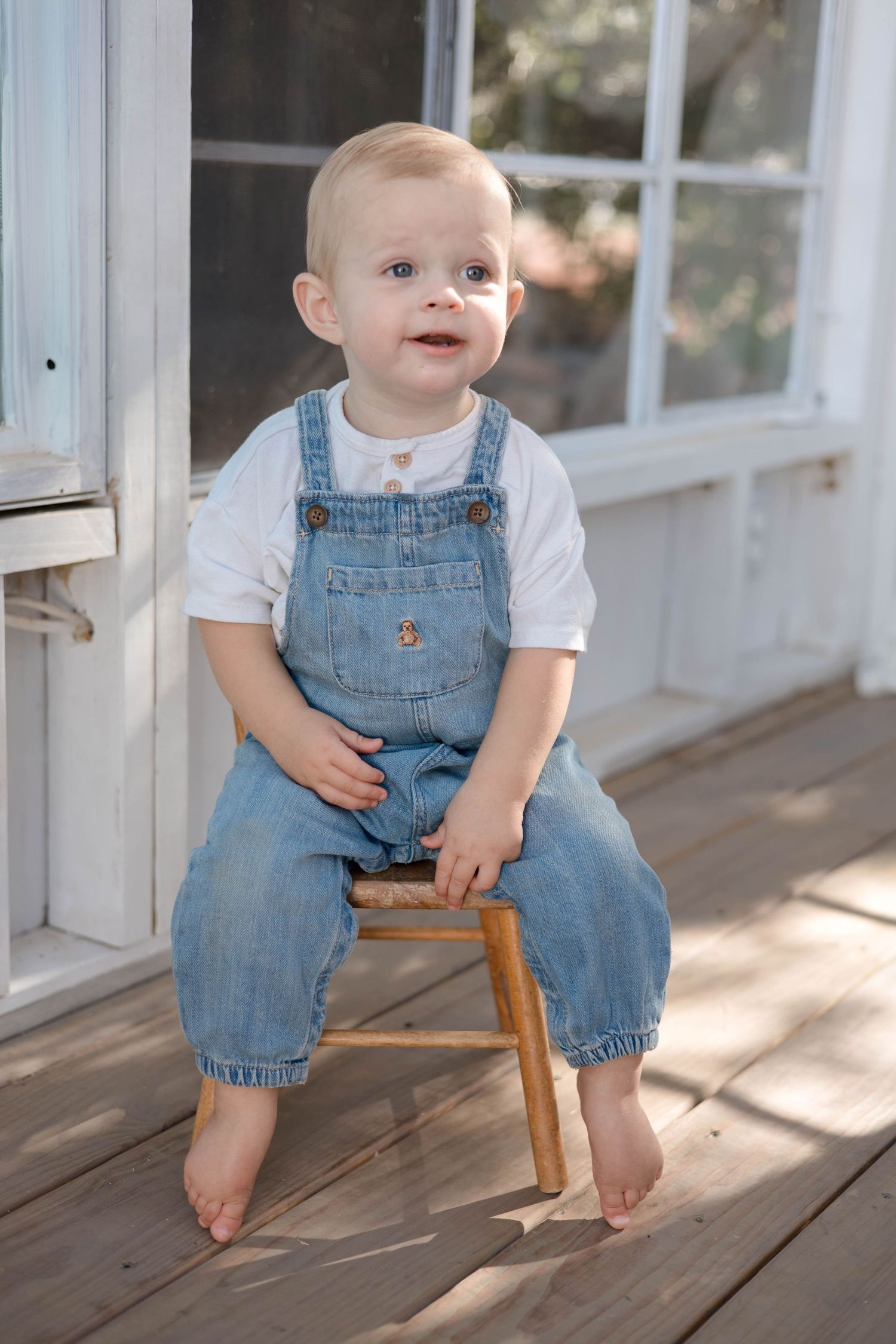 Baby wearing denim overalls and white shirt sits on wooden chair in sunlit porch setting.