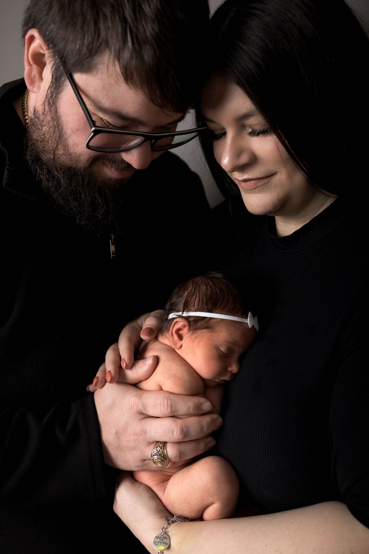 New parents in black clothing tenderly hold their sleeping newborn baby girl wearing a white headband.