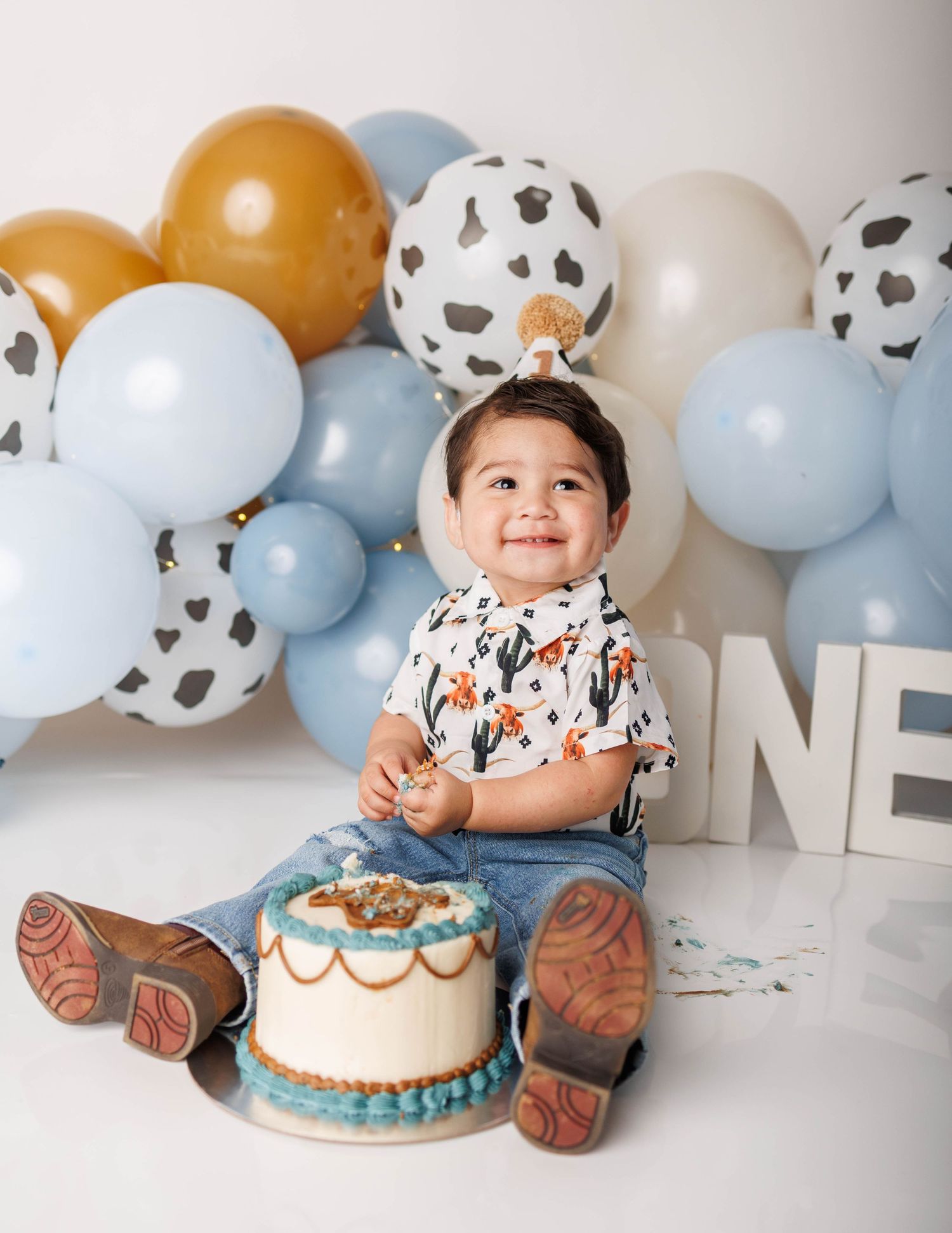 Baby boy smiling at his first birthday cake smash with western cowboy theme balloons and ONE sign.