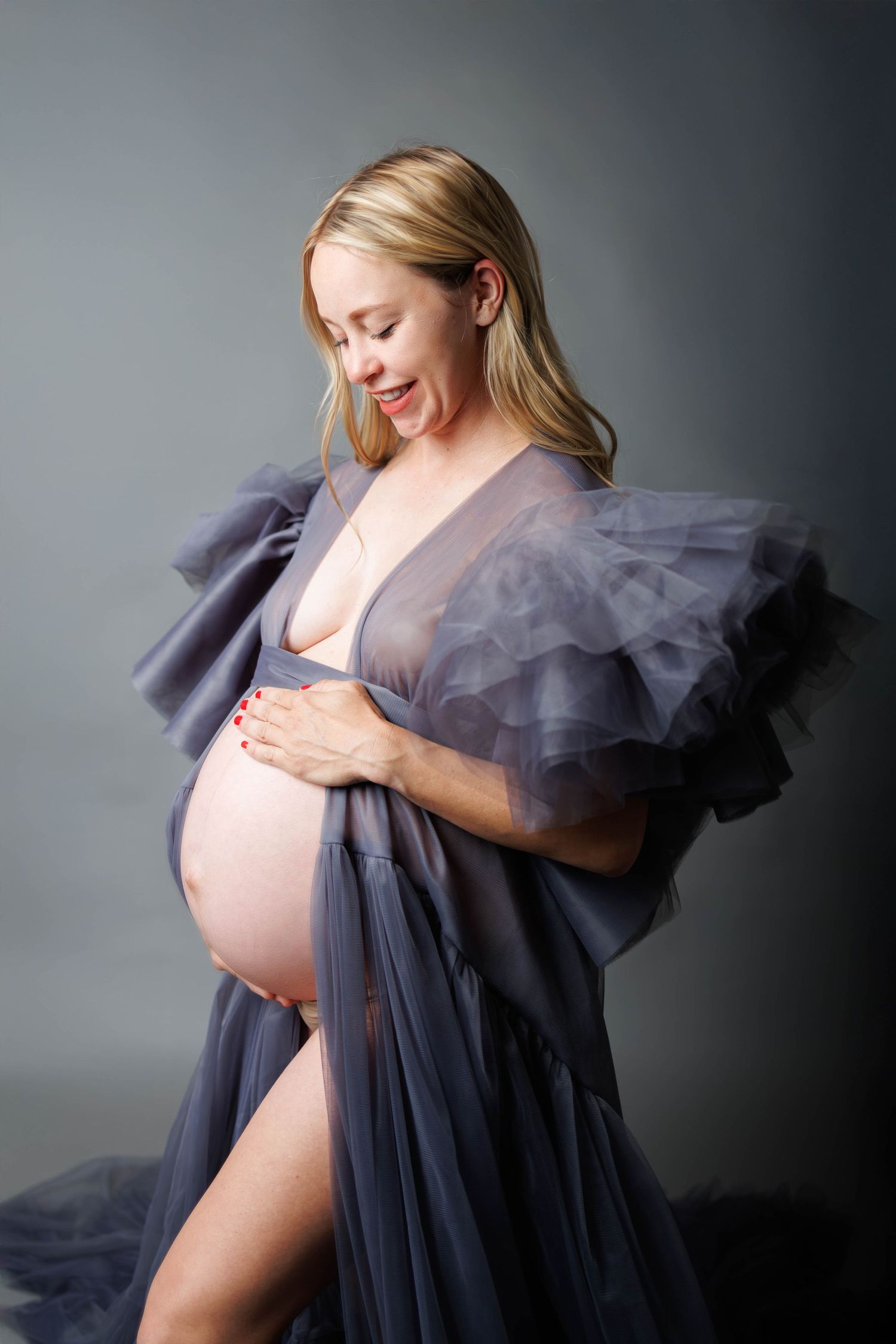 Smiling pregnant woman in sheer gray ruffled maternity robe cradling baby bump against gray studio background.