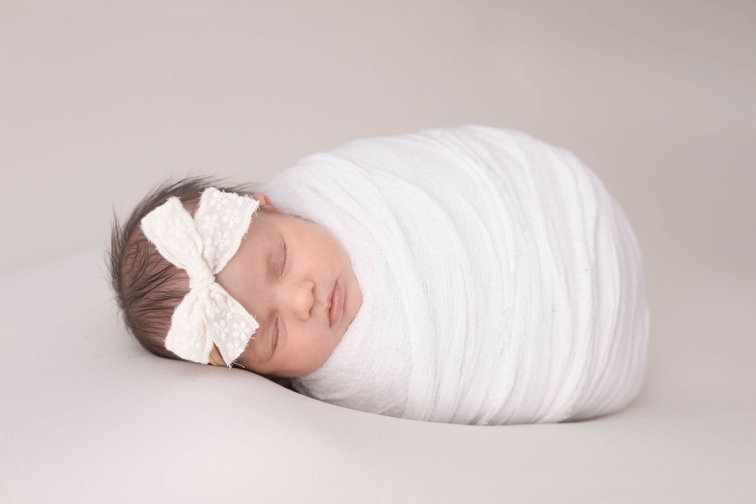 Sleeping newborn baby wrapped in white fabric with a lace bow headband on neutral background.