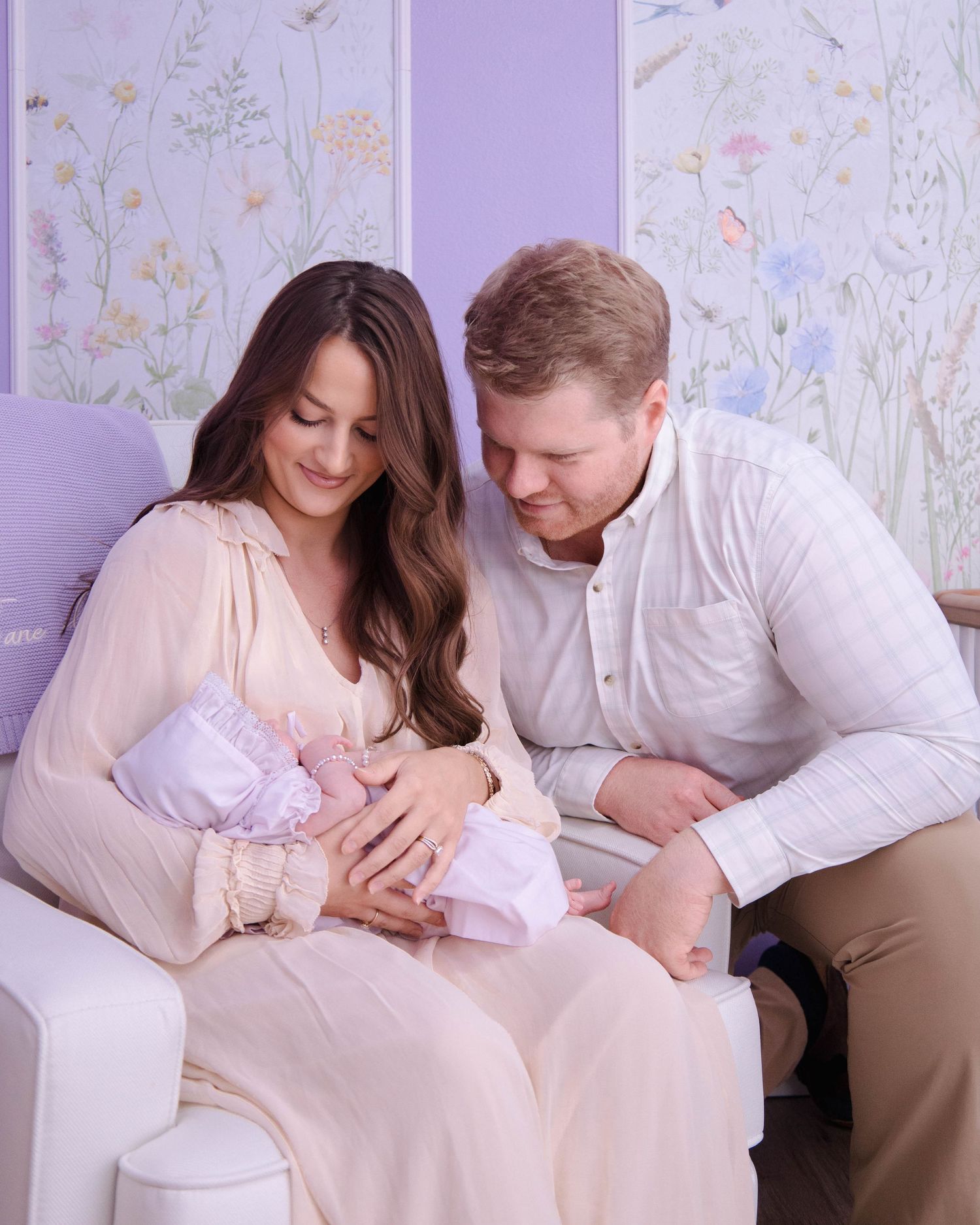 A couple in light-colored outfits sits together on a couch with a baby against a floral wallpapered purple wall.