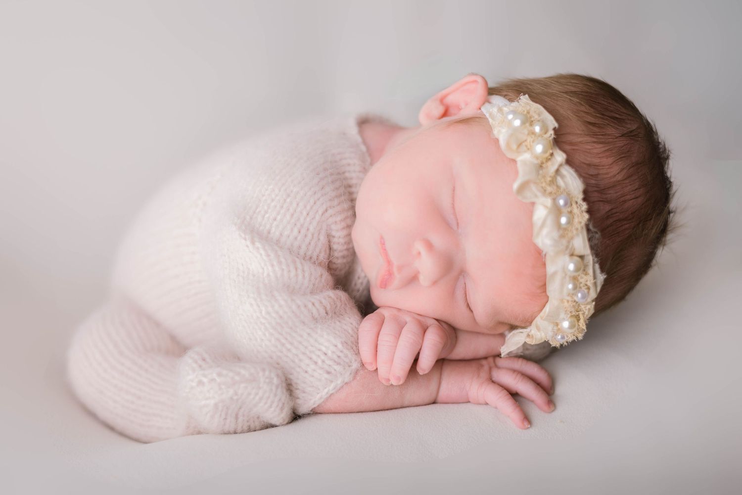 A sleeping newborn in pink clothing lies curled up on a soft gray backdrop.