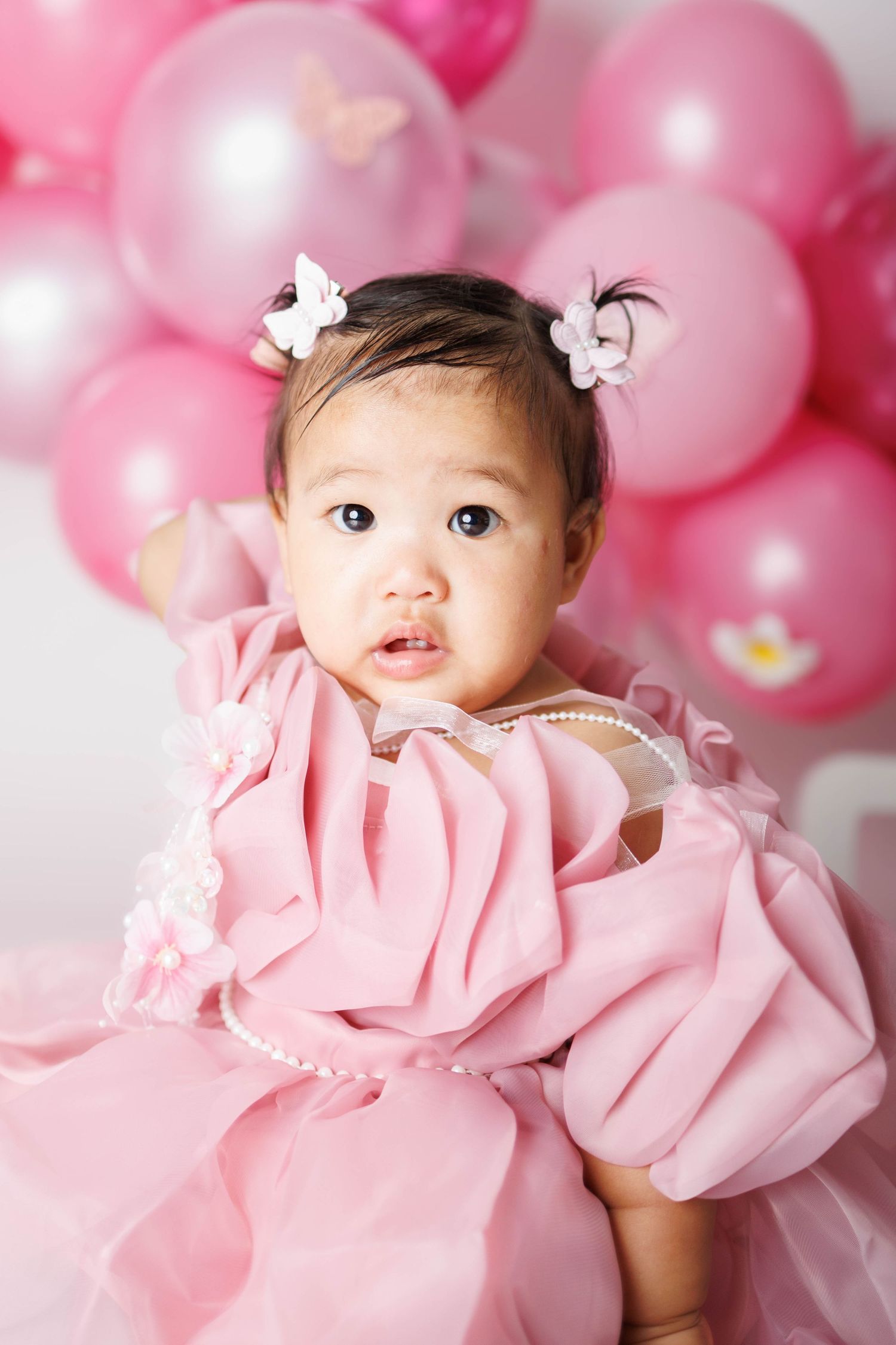 Adorable baby girl in a pink ruffled dress with butterfly hair clips, surrounded by pink balloons.