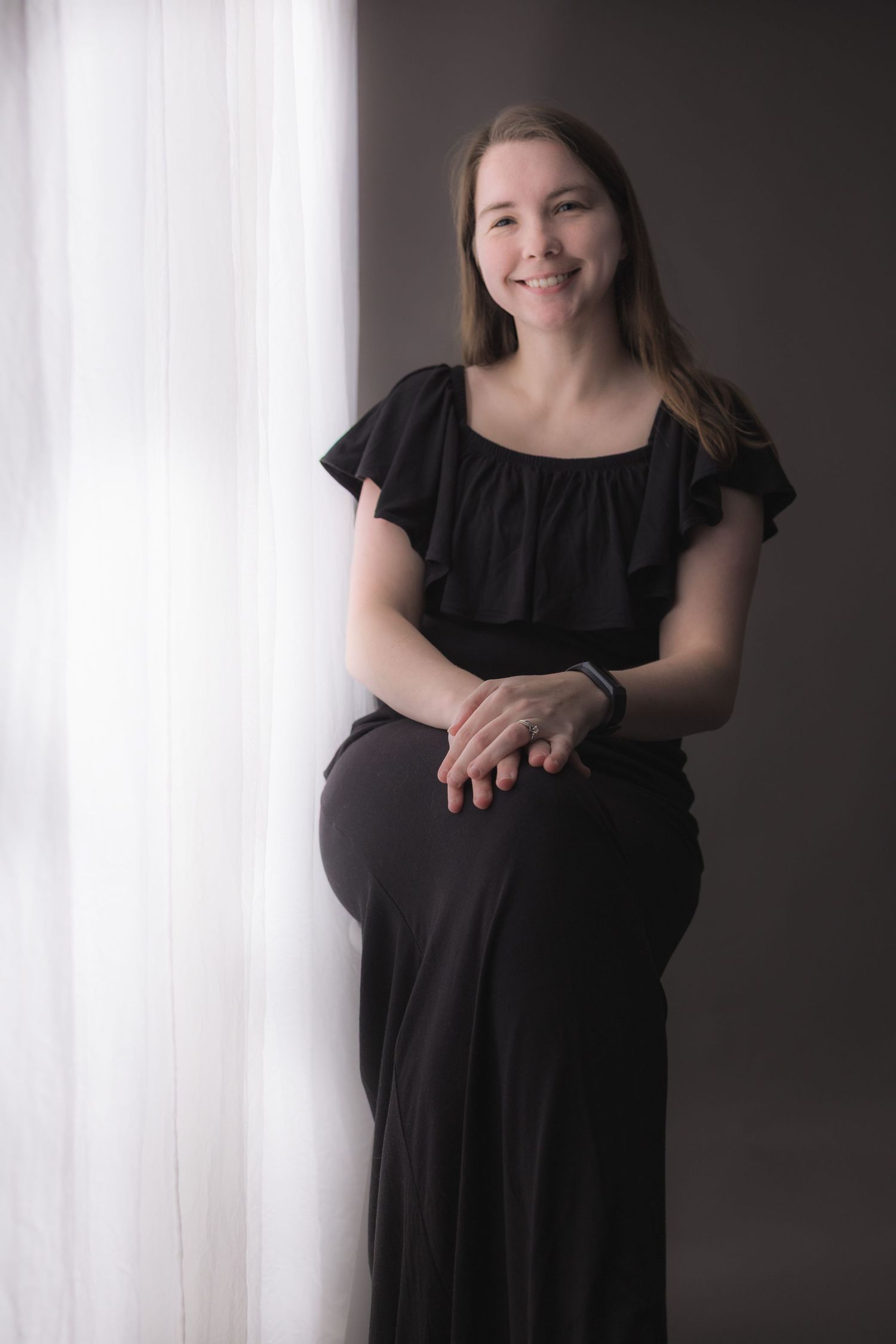 Smiling woman in black off-shoulder dress sits gracefully by a sheer white curtain against a gray background.