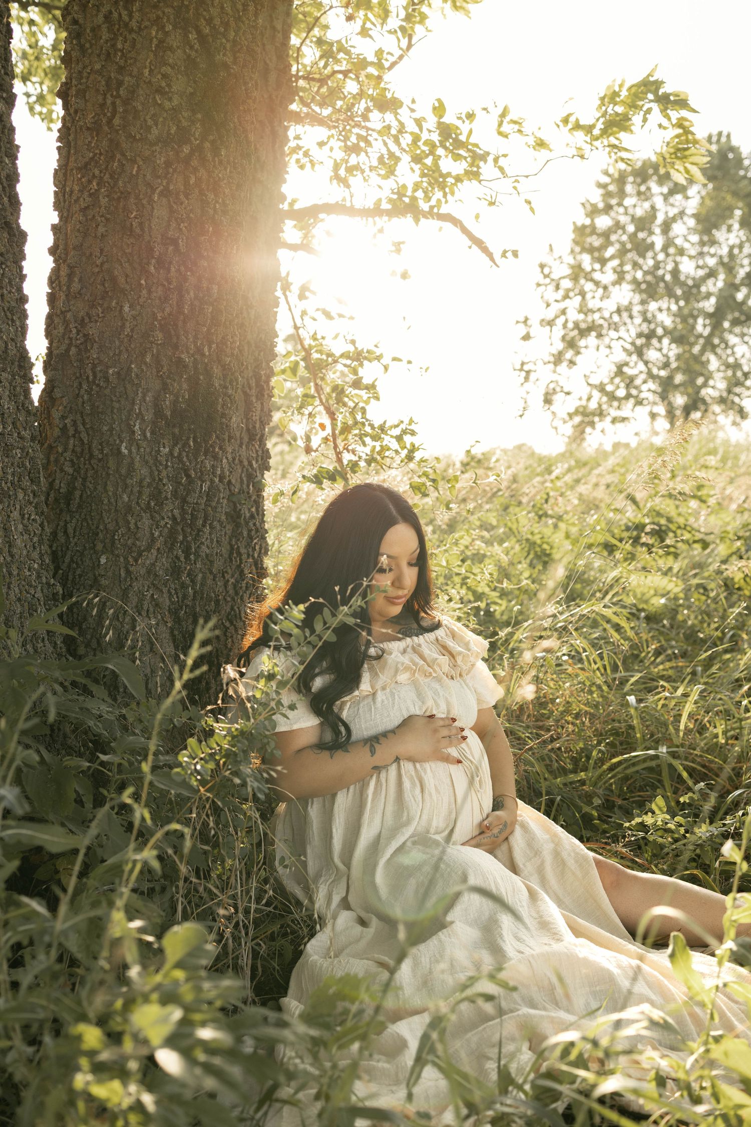 Pregnant woman in white dress sitting peacefully against a tree in a sunlit meadow, cradling her baby bump.