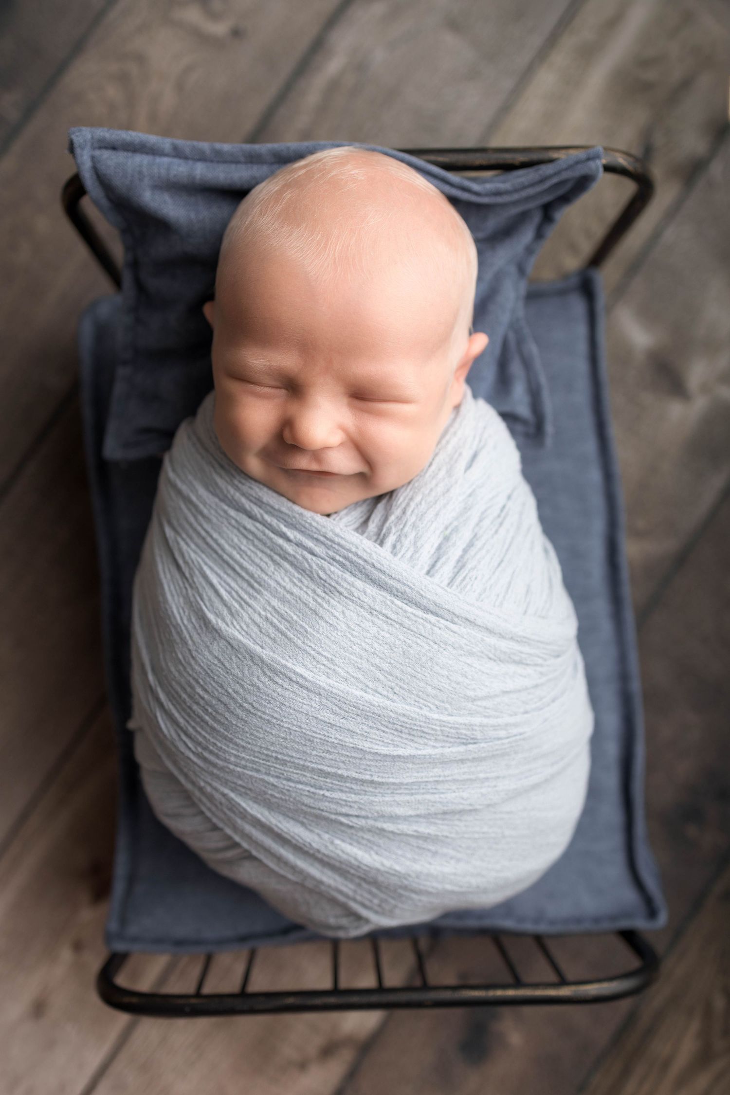 Smiling newborn baby swaddled in a light blue wrap, resting in a small metal bed on a wooden floor.