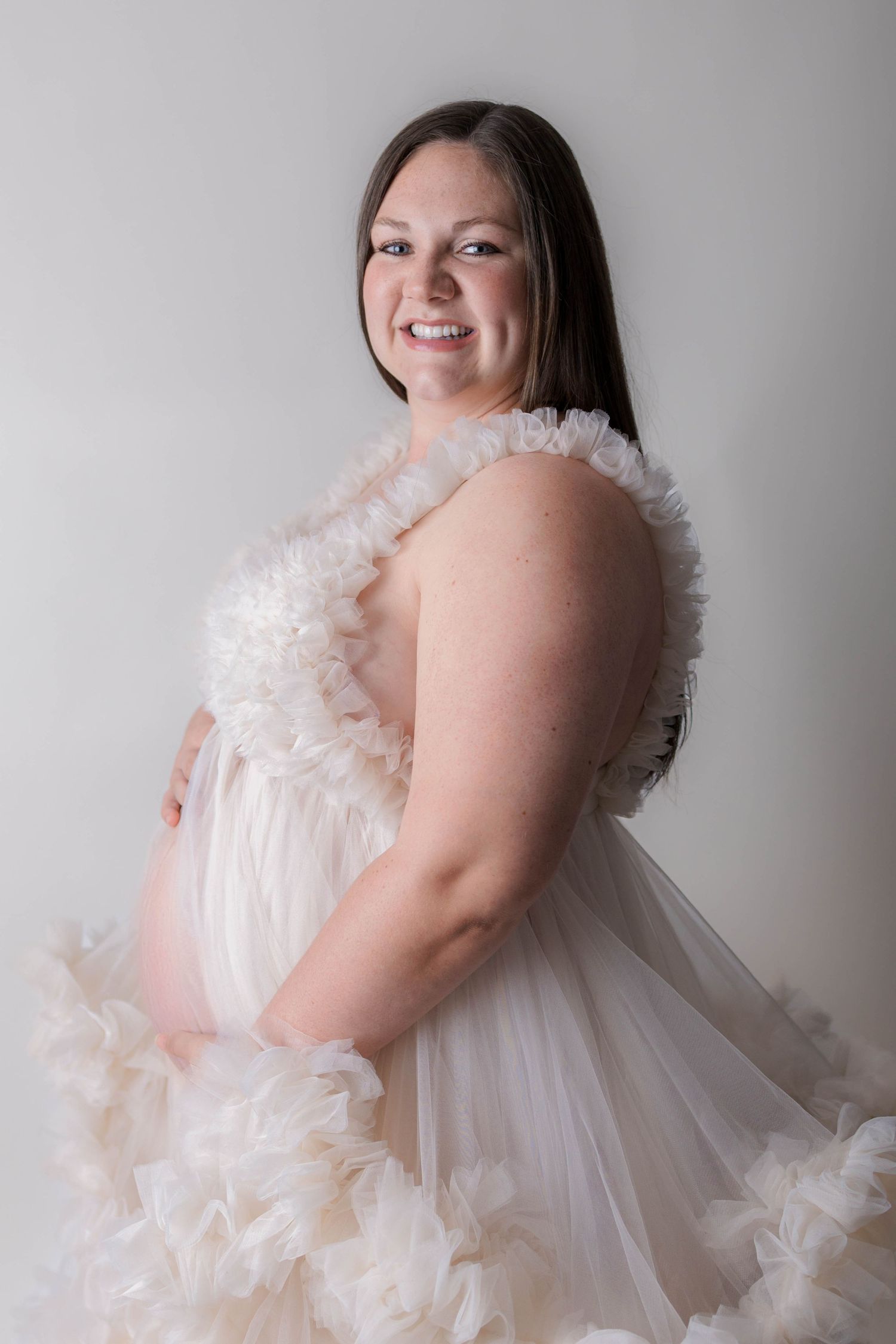 Smiling pregnant woman in a flowing white ruffled tulle maternity gown posing in a studio portrait.