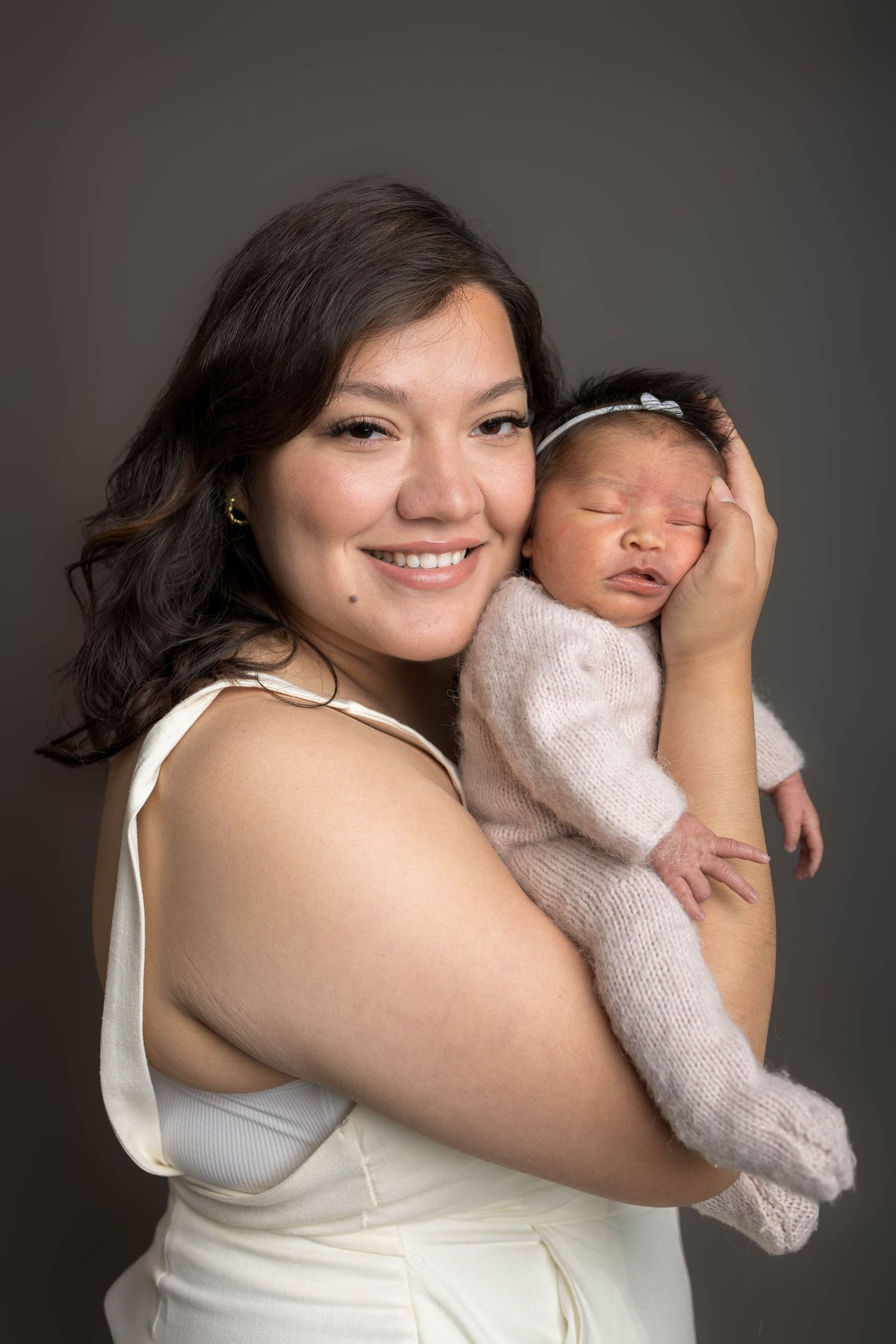 Smiling young mother in white dress holds sleeping newborn baby in pink knit outfit against gray studio background.
