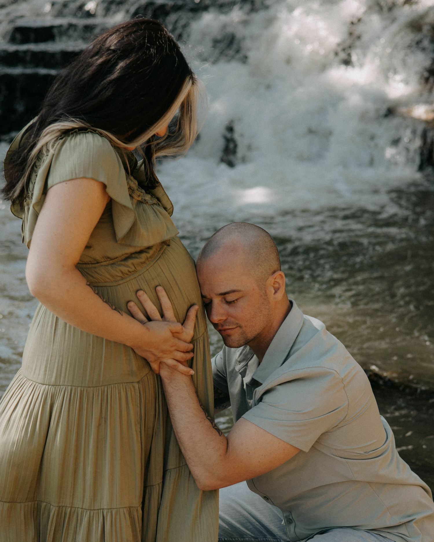 A tender maternity moment captured in front of a rushing waterfall with morning sunlight.
