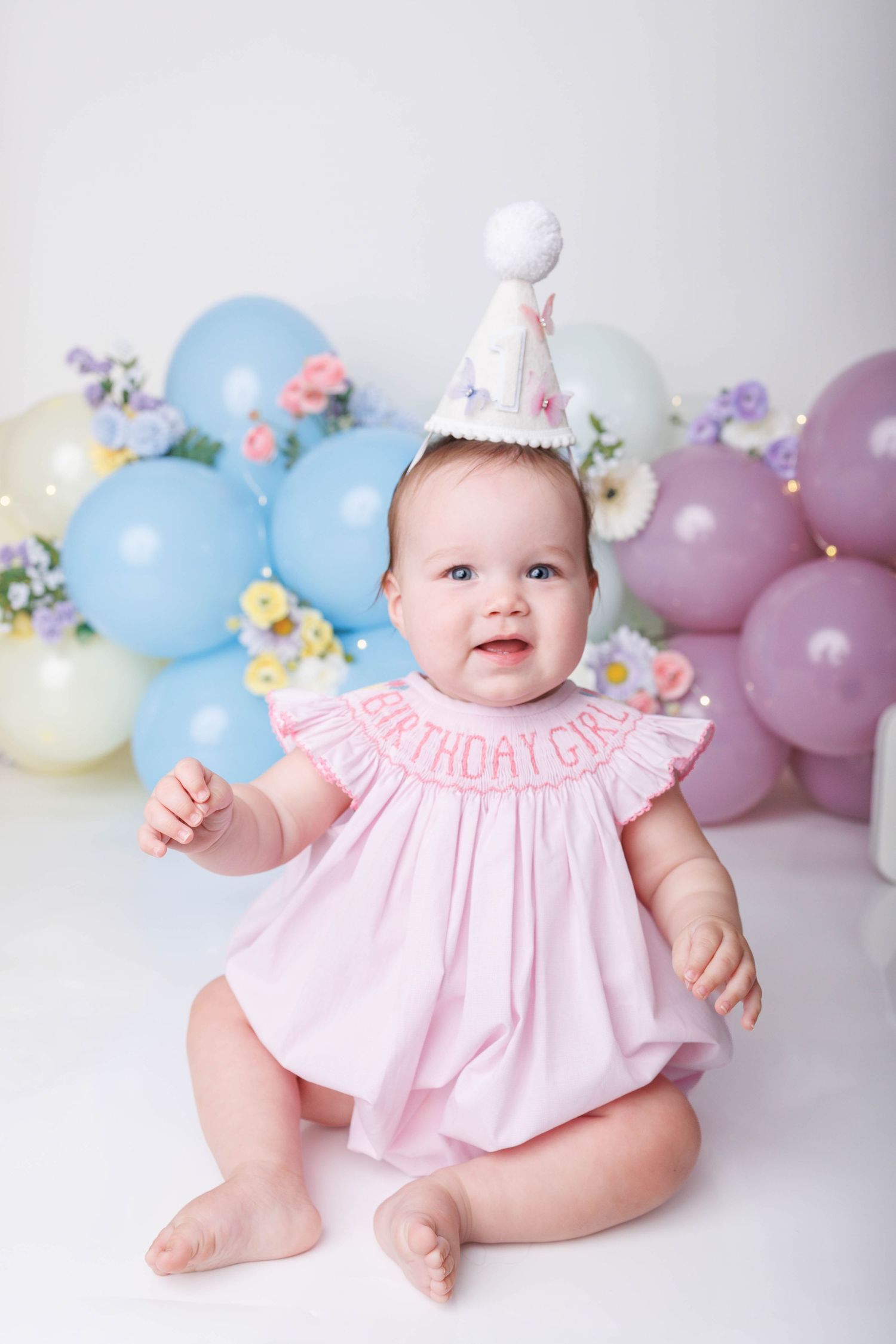 Baby girl in pink birthday dress and party hat sits surrounded by pastel balloons for her first birthday photo.