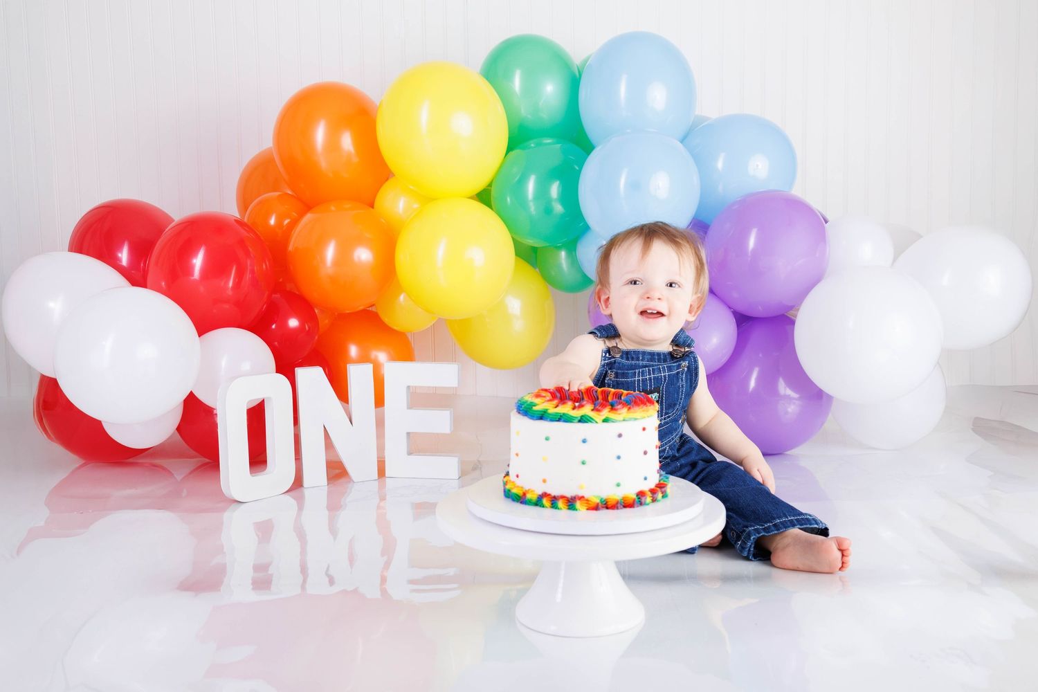 First birthday celebration setup with rainbow balloons, white ONE letters, and a colorful birthday cake on white backdrop.