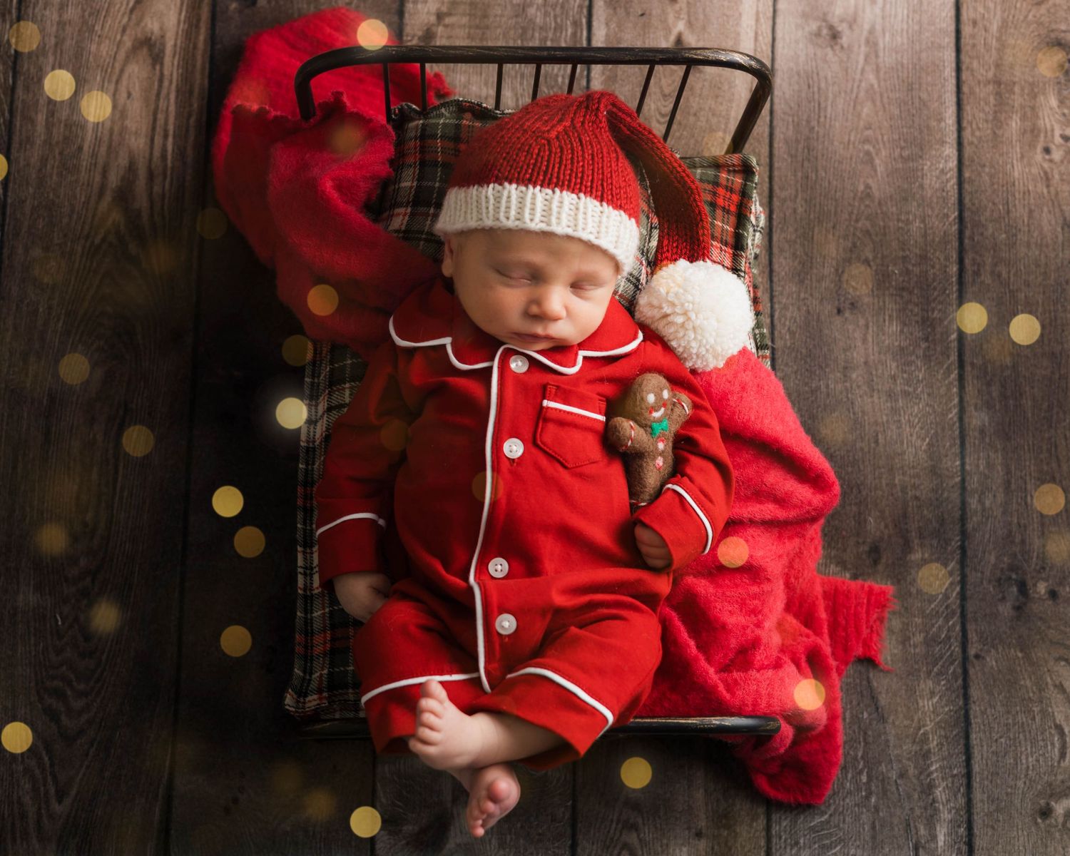 A baby in red holiday pajamas sits against a rustic wooden backdrop with twinkling lights.