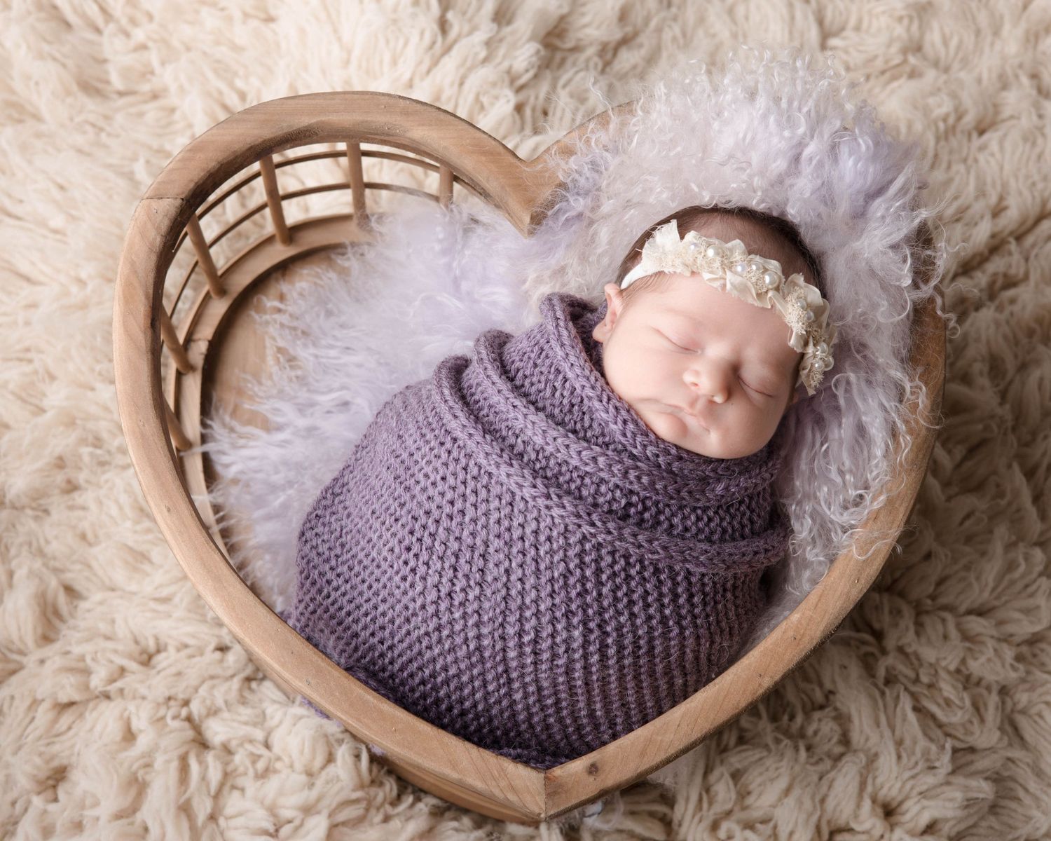 Newborn in purple wrap sleeping in heart-shaped basket on fuzzy backdrop.