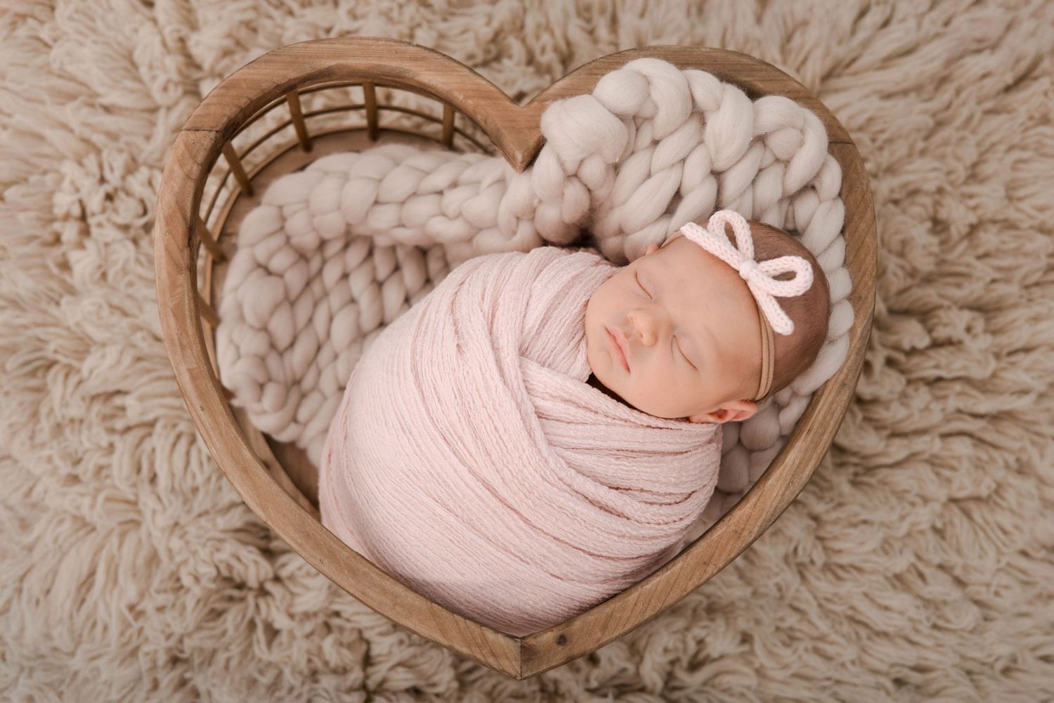 A sleeping newborn swaddled in soft pink fabric lies peacefully in a wooden heart-shaped bowl on fluffy beige carpet.