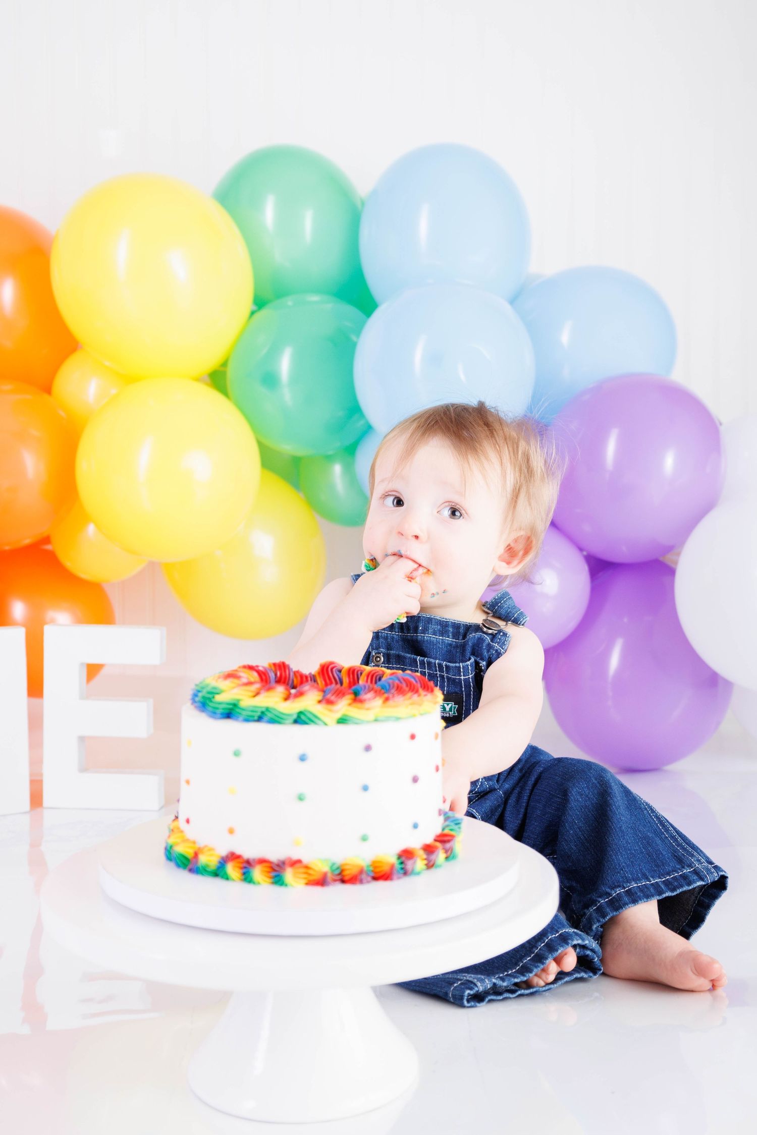 Colorful birthday cake on white stand with rainbow balloon arch backdrop for first birthday celebration.