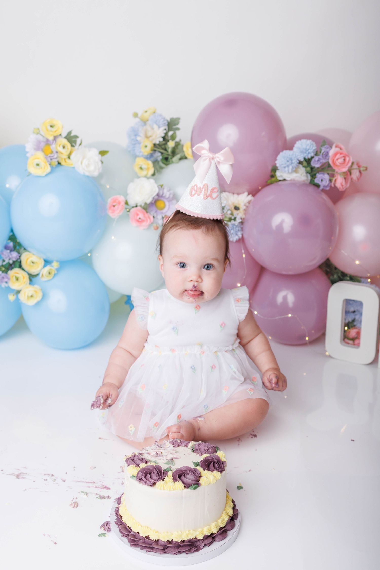 Baby girl in white dress with cake smash birthday setup featuring blue and purple balloons and floral decorations.